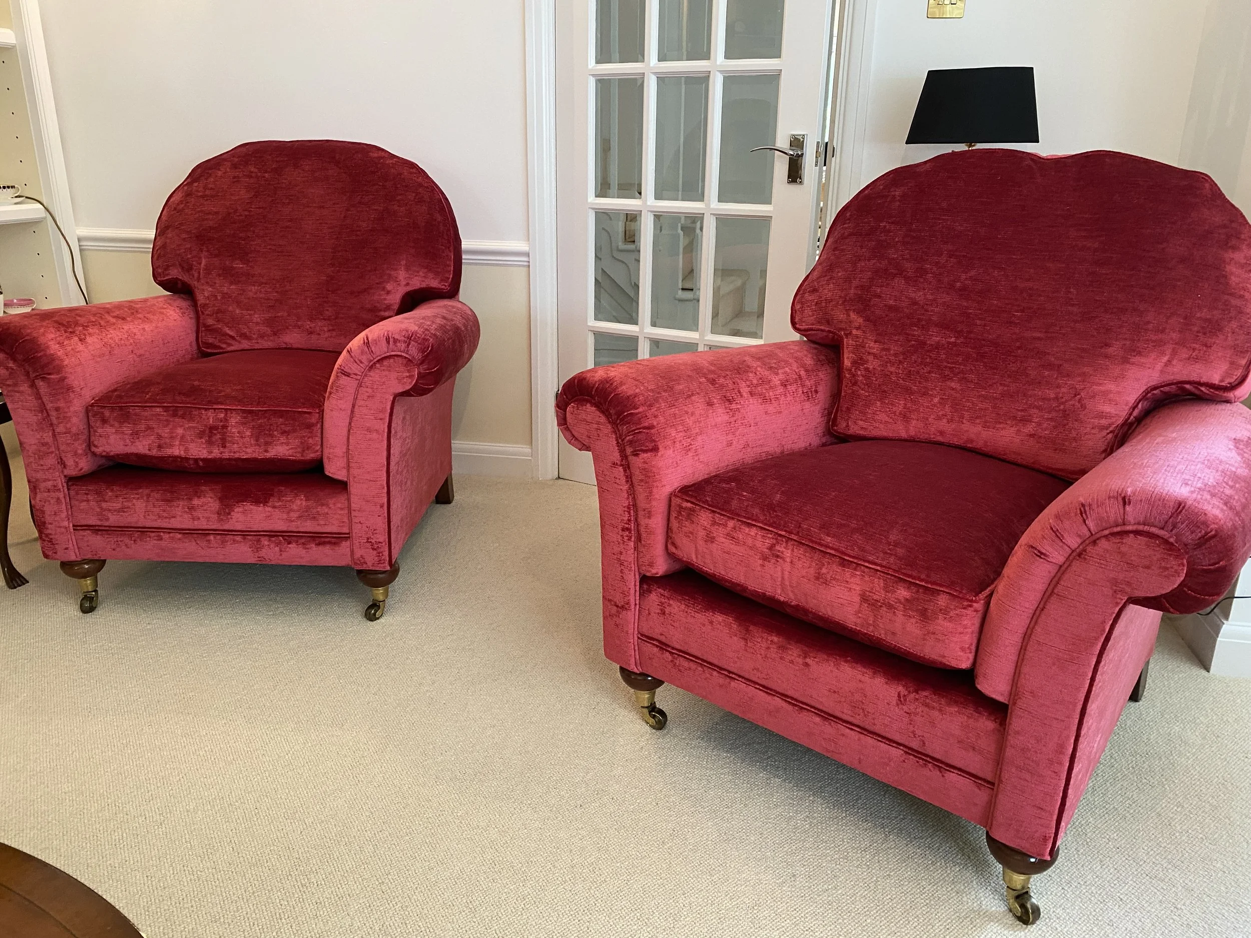 Two red velvet armchairs with wooden legs and brass casters are positioned on a beige carpet in a room with white walls. A glass-paneled door and a black table lamp are visible in the background.