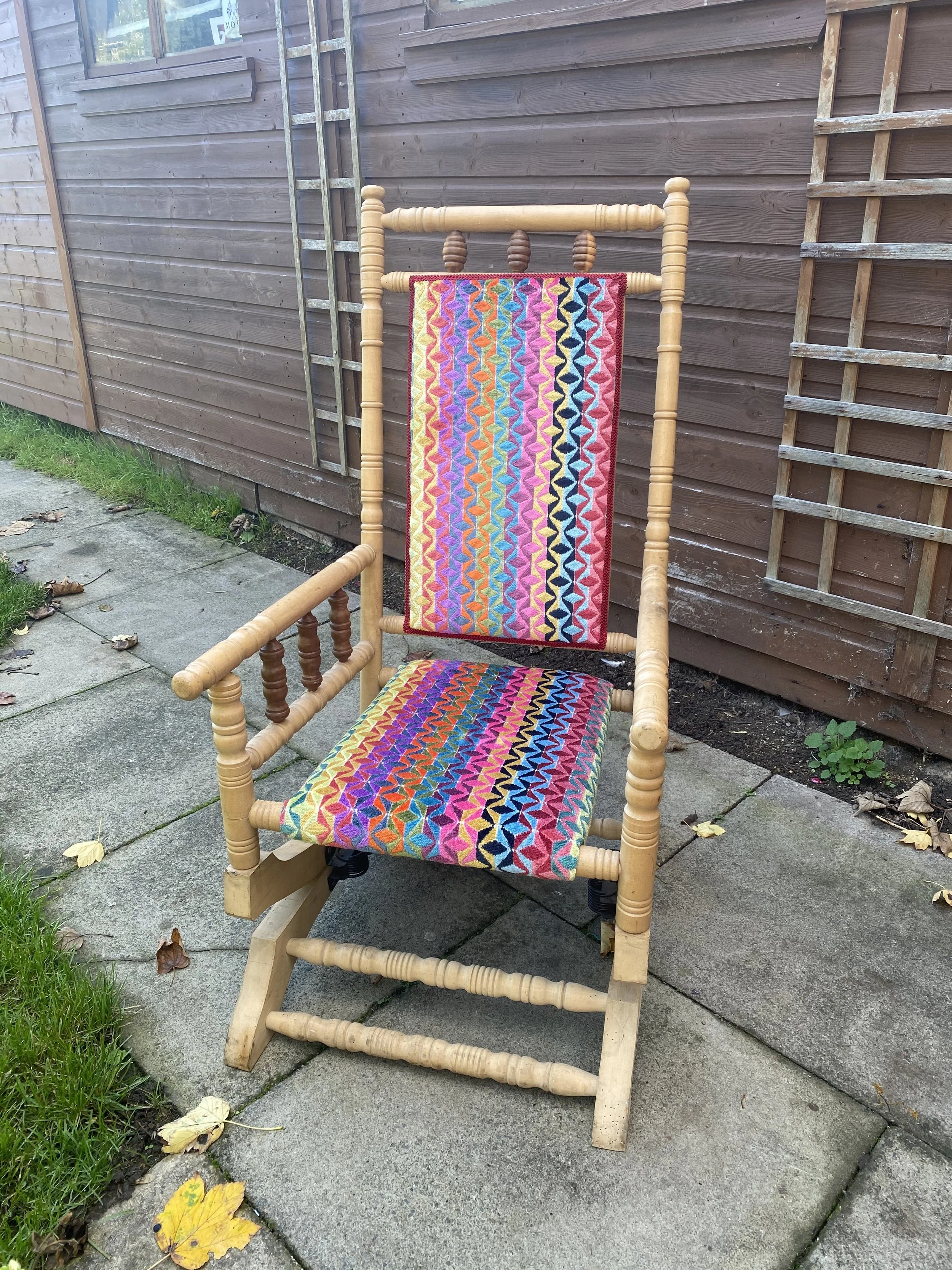 A wooden rocking chair with colorful, embroidered fabric cushions on the seat and backrest, placed outdoors on a paved area next to a brown wooden fence.