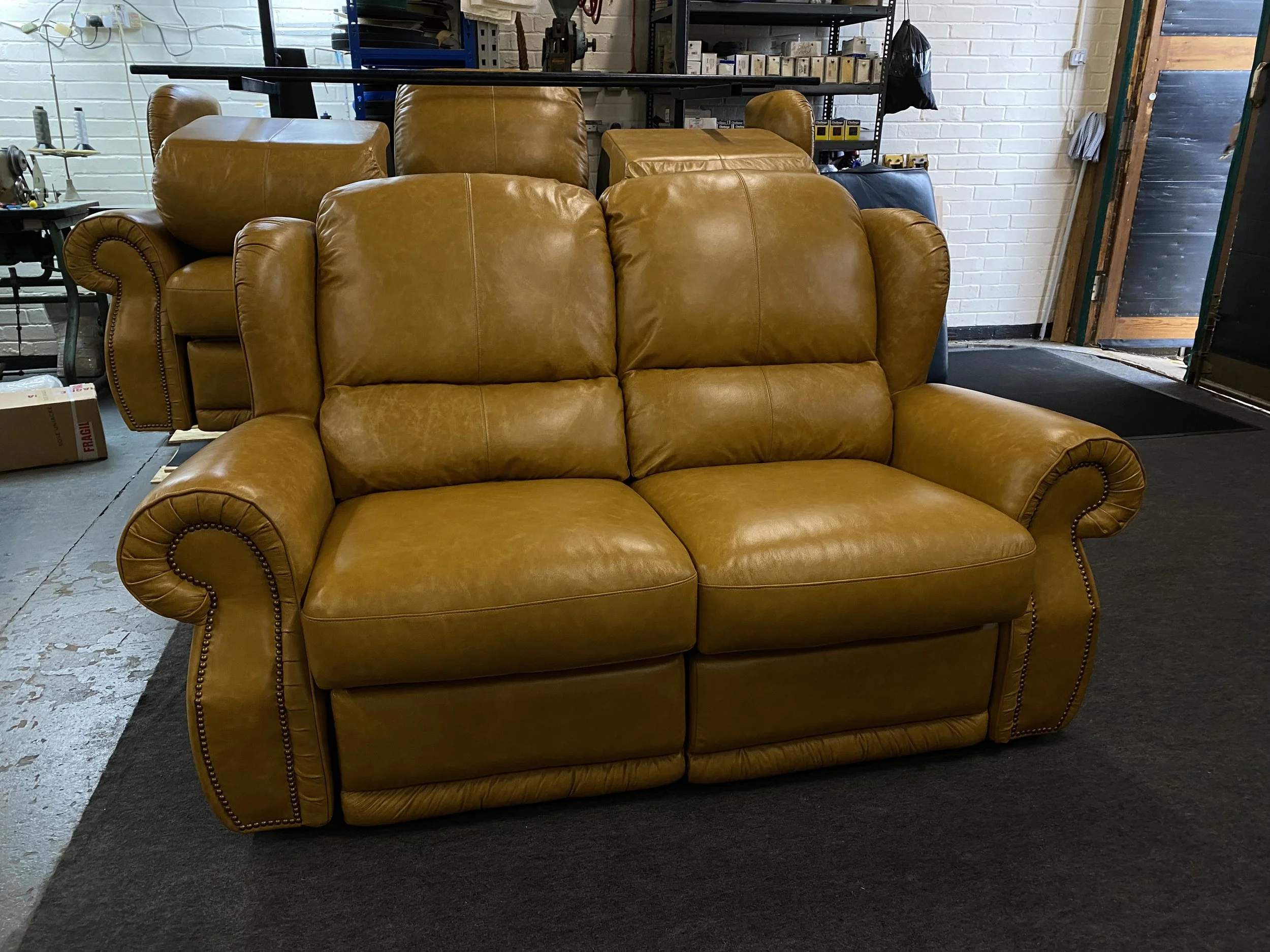 A tan leather recliner sofa with rolled arms and decorative nailhead trim, placed on a dark carpet in a furniture store or warehouse.