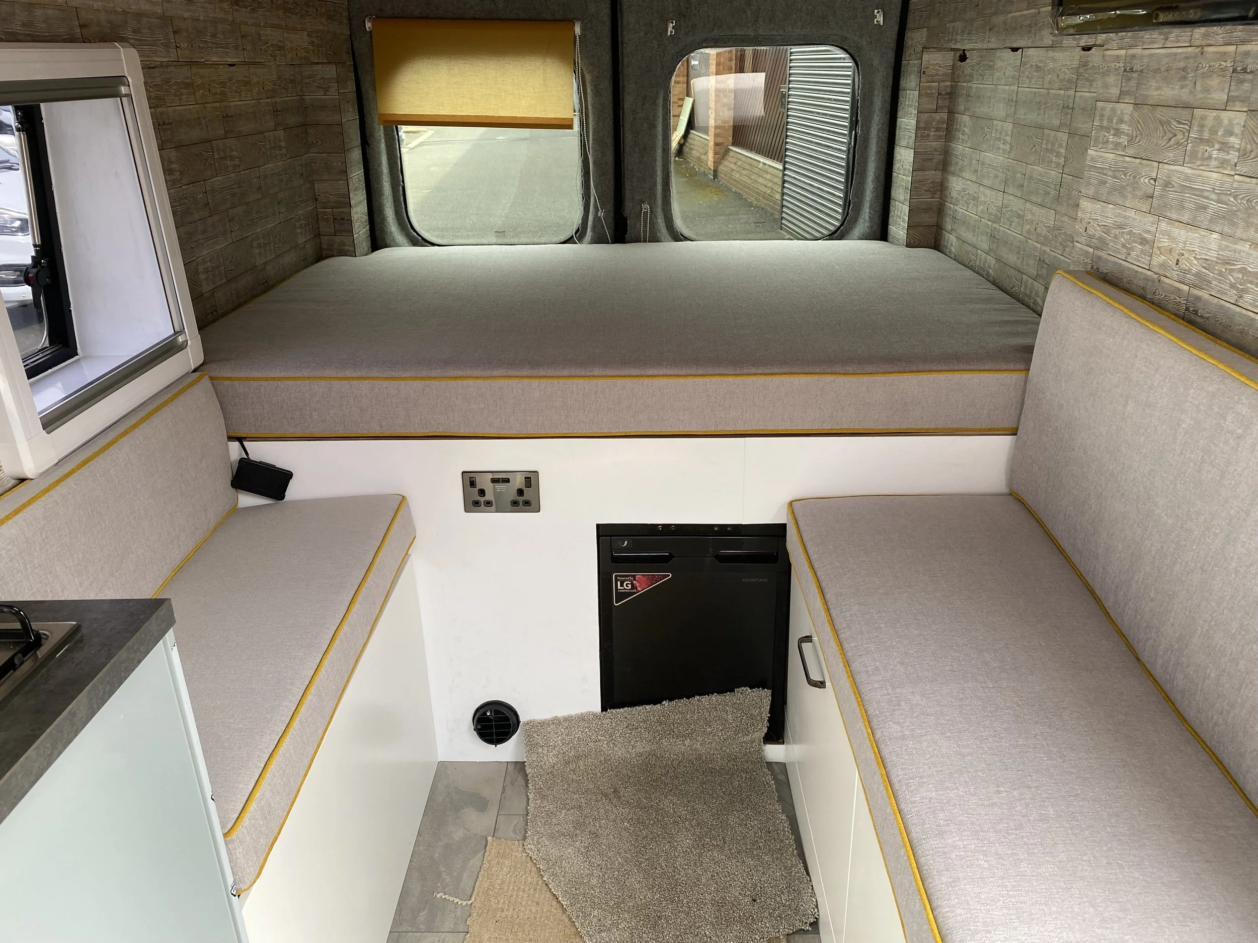 Interior of a camper van with a built-in bed on the upper level, two cushioned seating areas below, and a small refrigerator. The van has wood-paneled walls, a window with a rolled-up blind, electrical outlets, and a small carpet on the floor.