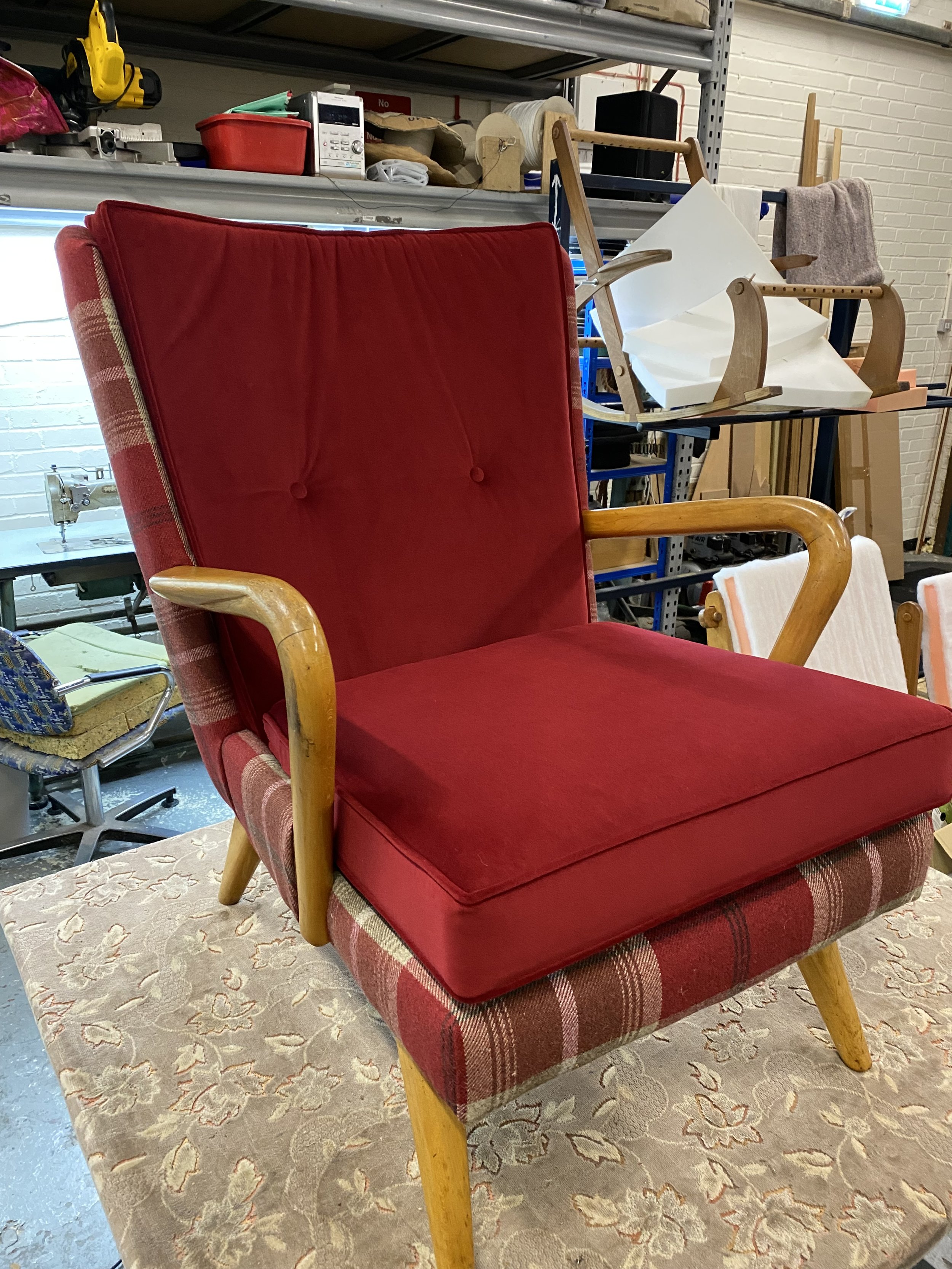 Red upholstered armchair with wooden arms and plaid fabric on the sides, placed on a floral-patterned rug in a workshop or storage area.