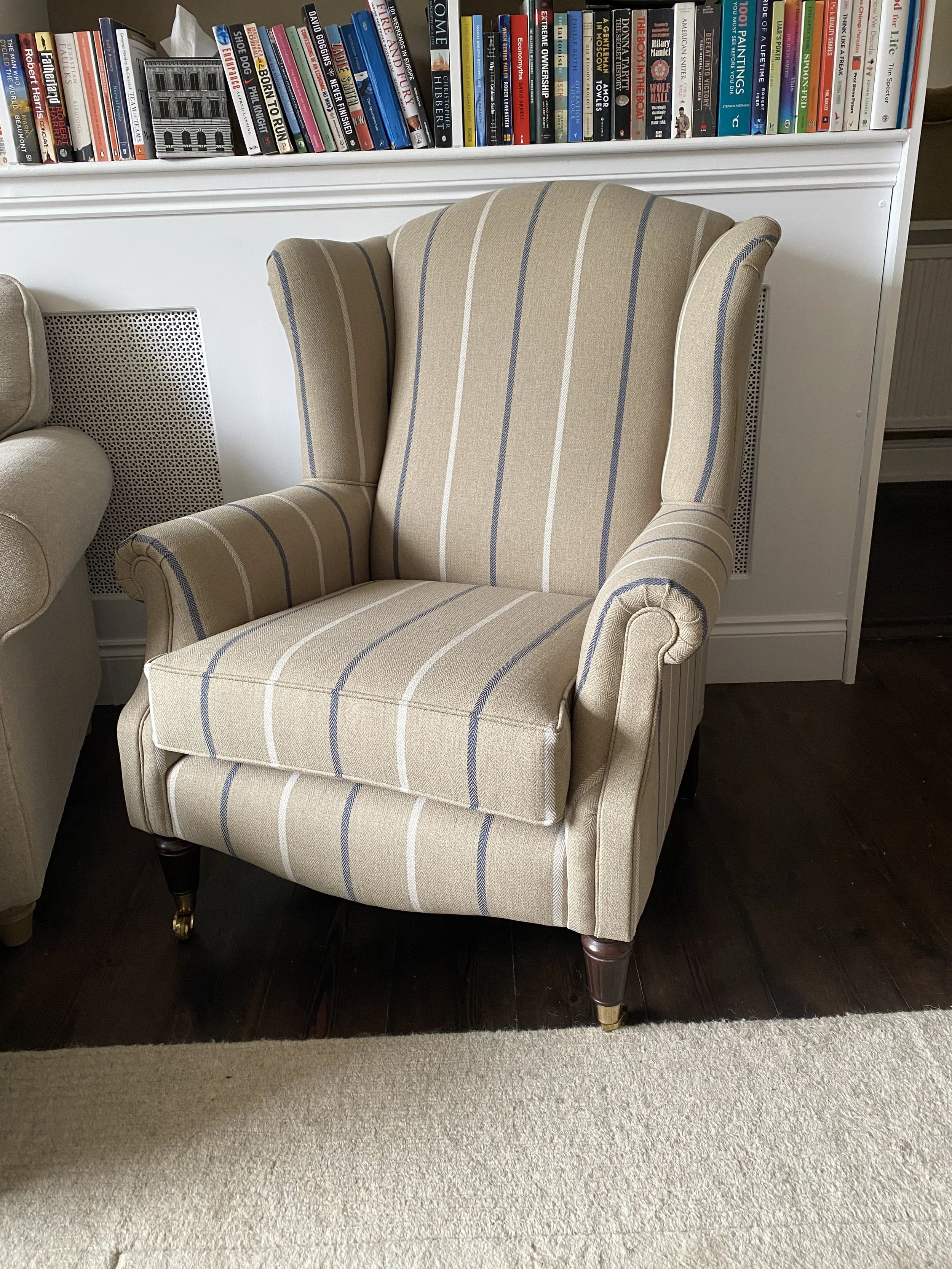 A beige armchair with vertical blue and white stripes, wooden legs with small brass caps, placed on a dark wooden floor next to a beige carpet, with a white bookshelf filled with colorful books in the background.