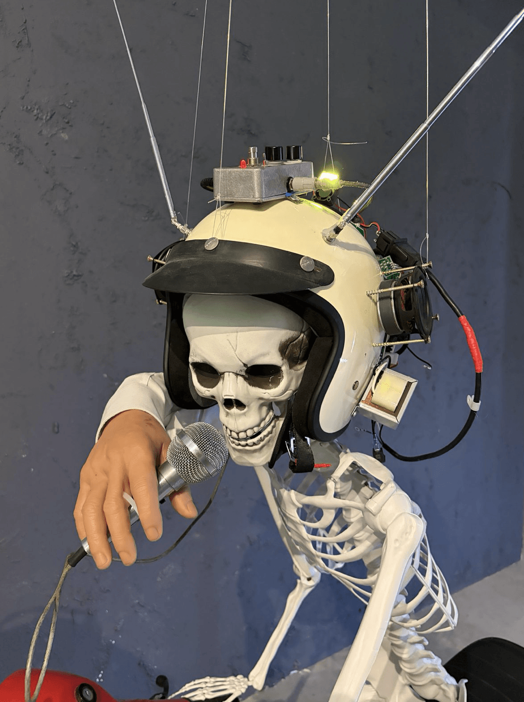 Detail. Skeleton figure in a modified motorcycle helmet fitted with electronics, speaker, and antennas, holding a microphone in a mannequin hand.