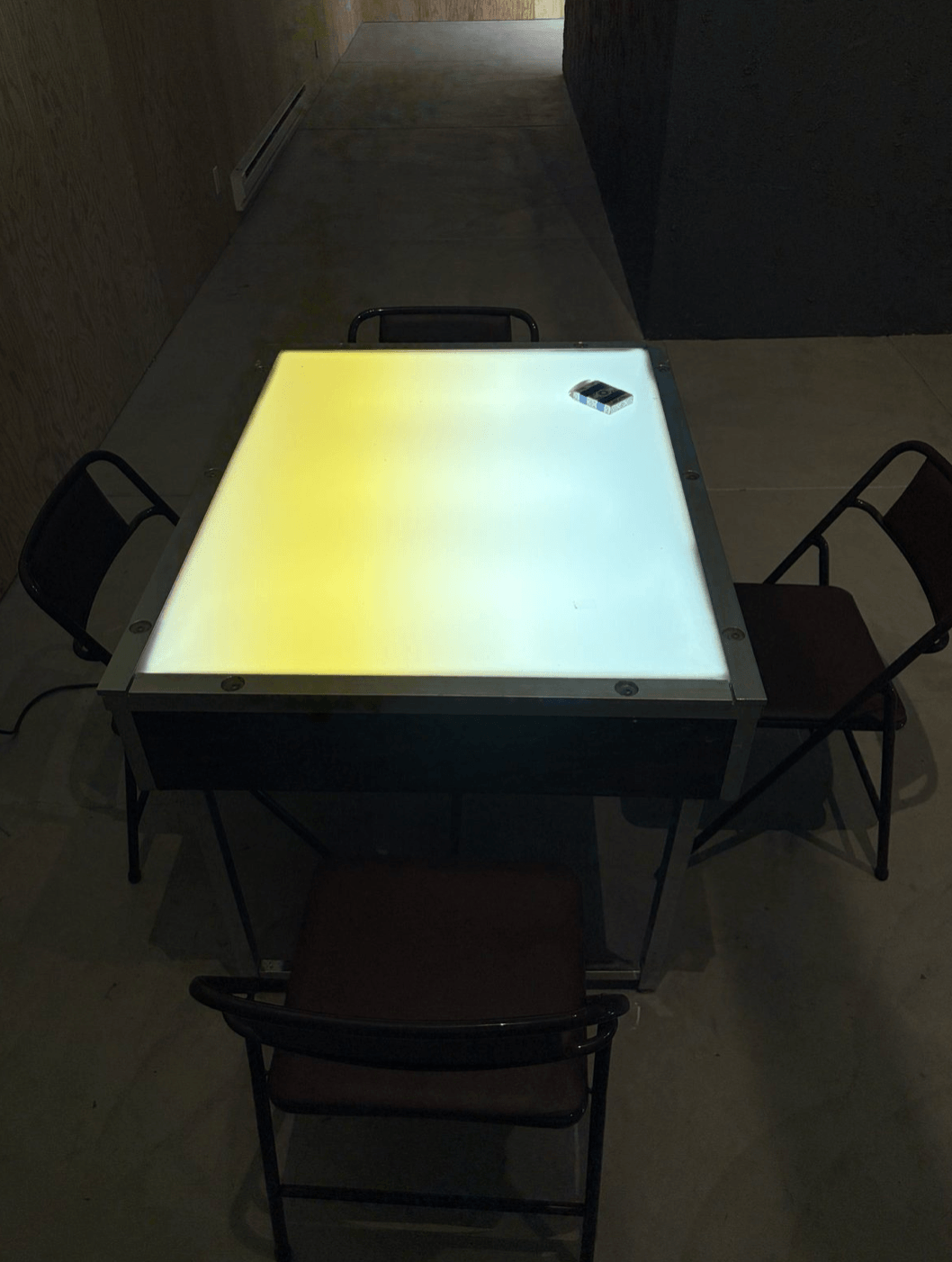 Backlit card table with folding chairs, opposite the entrance in the darkened main gallery.