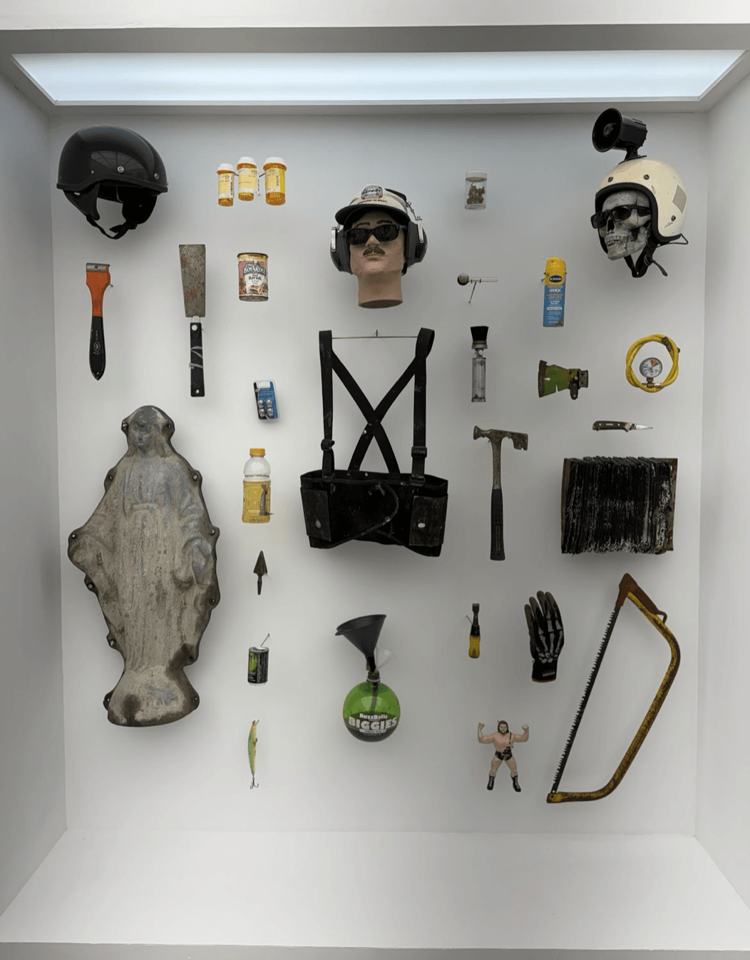  Inset display “The “Freelancer” Life”. Float-mounted objects including a cast Virgin Mary, a Gatorade bottle of yellow liquid, a rusted Chef Boyardee can, a BuzzBallz “Biggies,” prescription bottles, hand tools, and a skull in a helmet. 