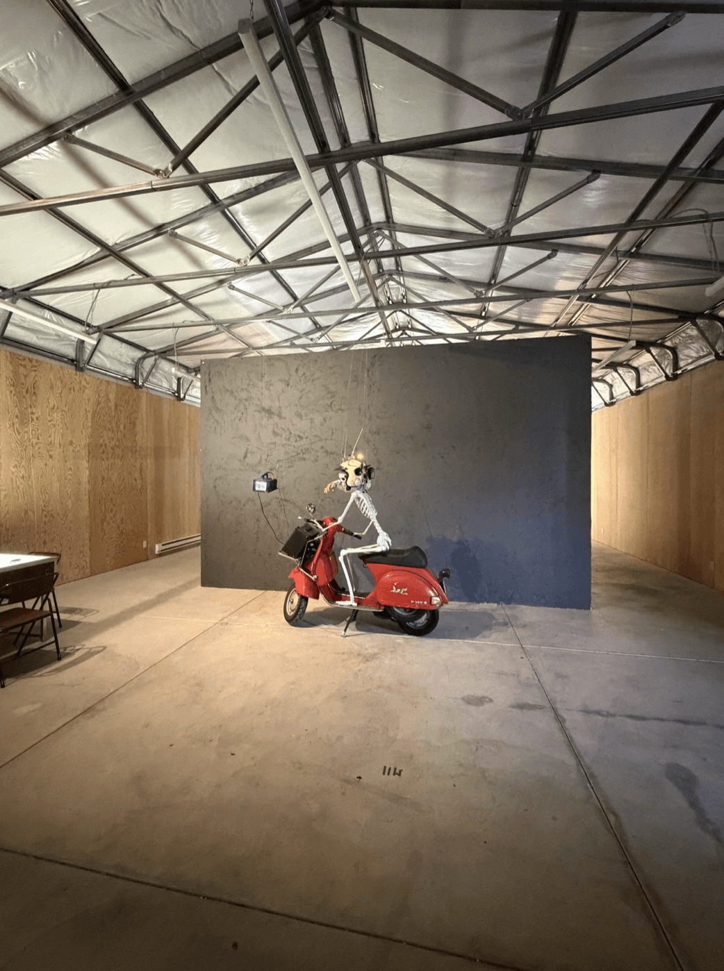 Installation view. The constructed gallery, painted dark gray, occupies roughly half the main gallery floor. Skeleton on Vespa and suspended CRT monitor at the entrance; backlit card table at left.