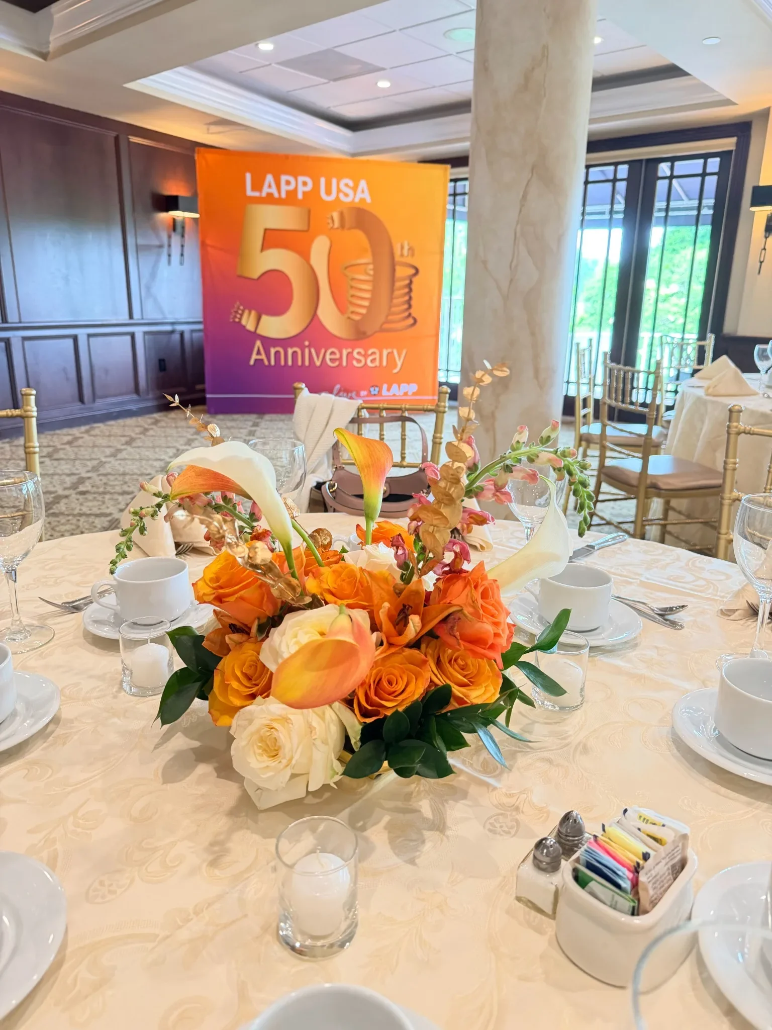 A banquet table with a floral centerpiece, plates, cups, utensils, and sugar packets, set for a celebration. In the background, a large orange sign commemorates LAPP USA's 50th anniversary with a stylized number 50 and a spool of wire illustration.