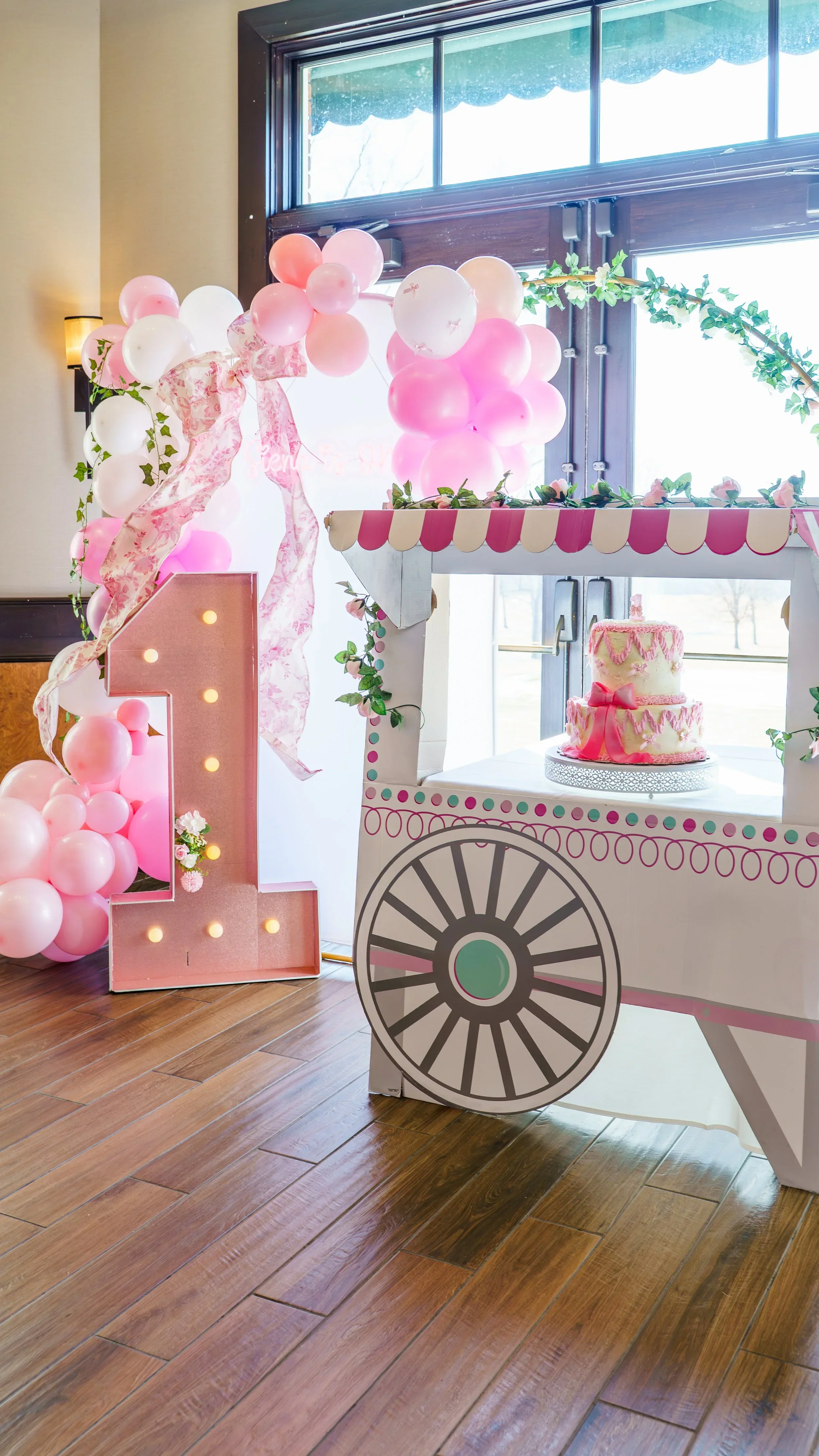 Pink and white balloons arranged in an arch next to a decorated pink cake on a white cart with a pink and white striped canopy, celebrating a first birthday.
