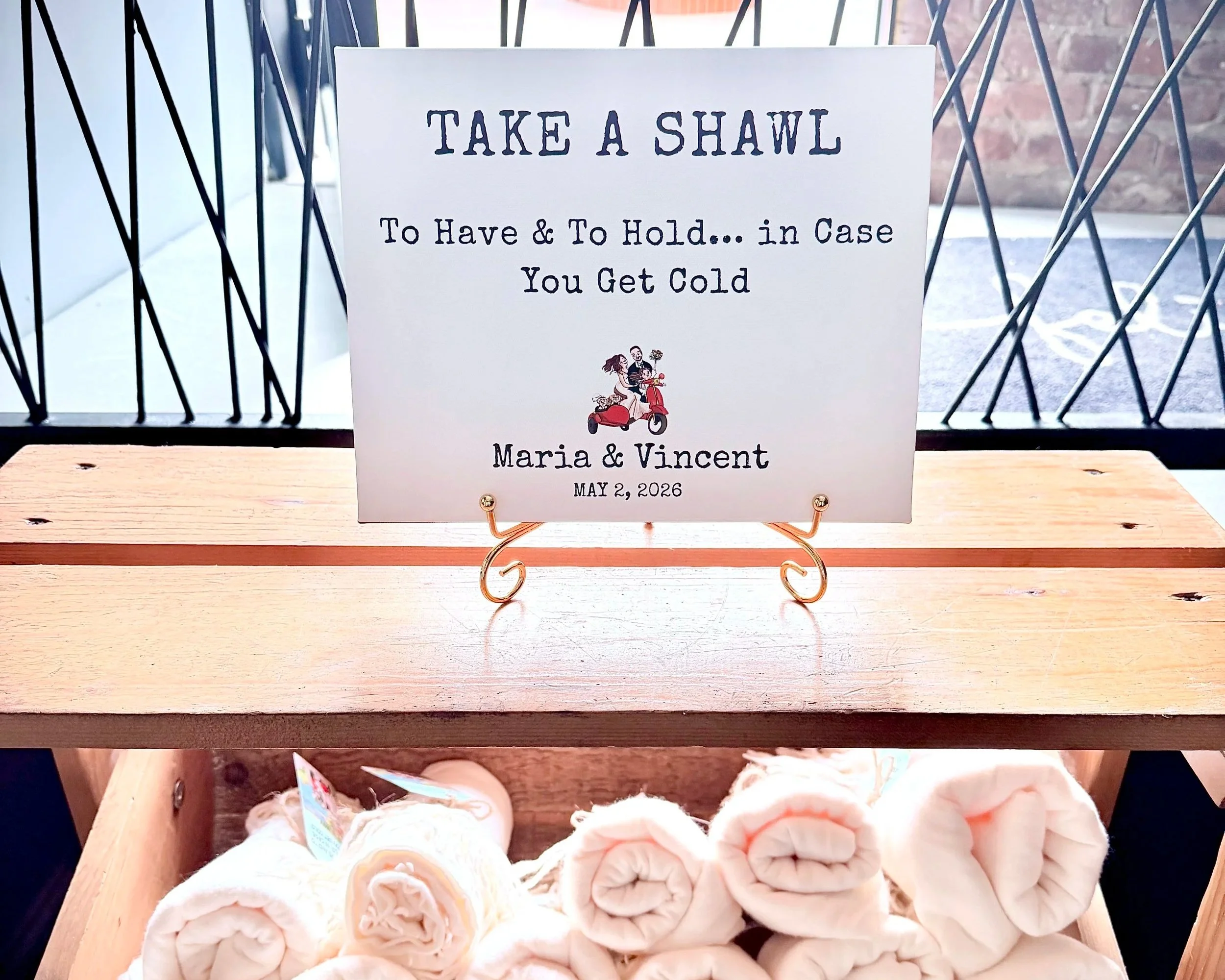A sign on a table that reads, 'Take a Shawl. To Have & To Hold... in Case You Get Cold. Maria & Vincent, May 2, 2026.' Below the sign are soft, fluffy, light-colored shawls or wraps arranged on the table.