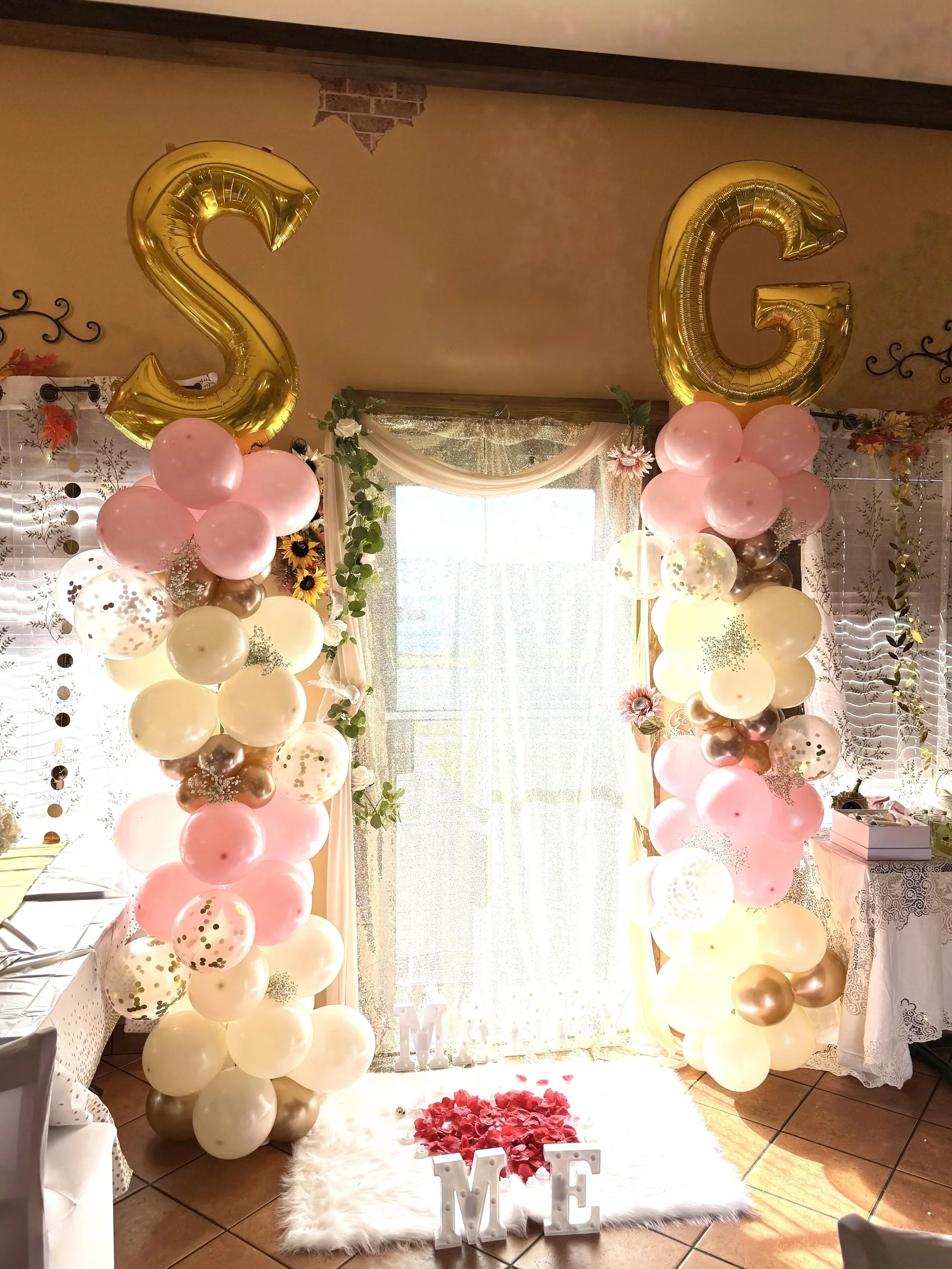 Decorative setup with large gold foil balloons forming the letters 'S' and 'G' above a floral balloon arch in pink, cream, gold, and white. The arrangement includes pink and gold balloons and features a white fluffy rug with red rose petals and the w