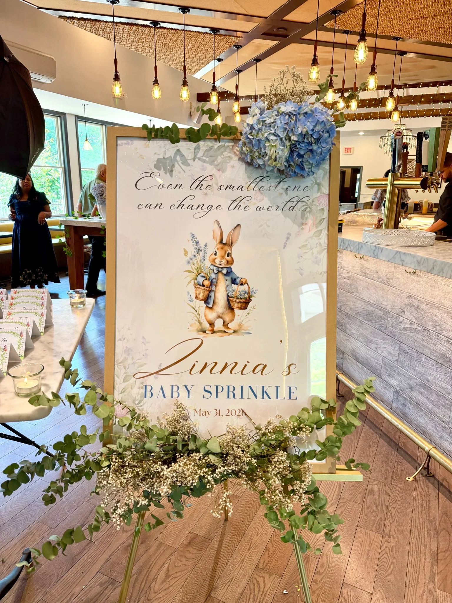 A decorative sign for a baby shower with the quote "Even the smallest one can change the world" and a picture of a rabbit holding a basket. The sign features floral and greenery accents, with a blue hydrangea flower. The event is for "Linnia's Baby S