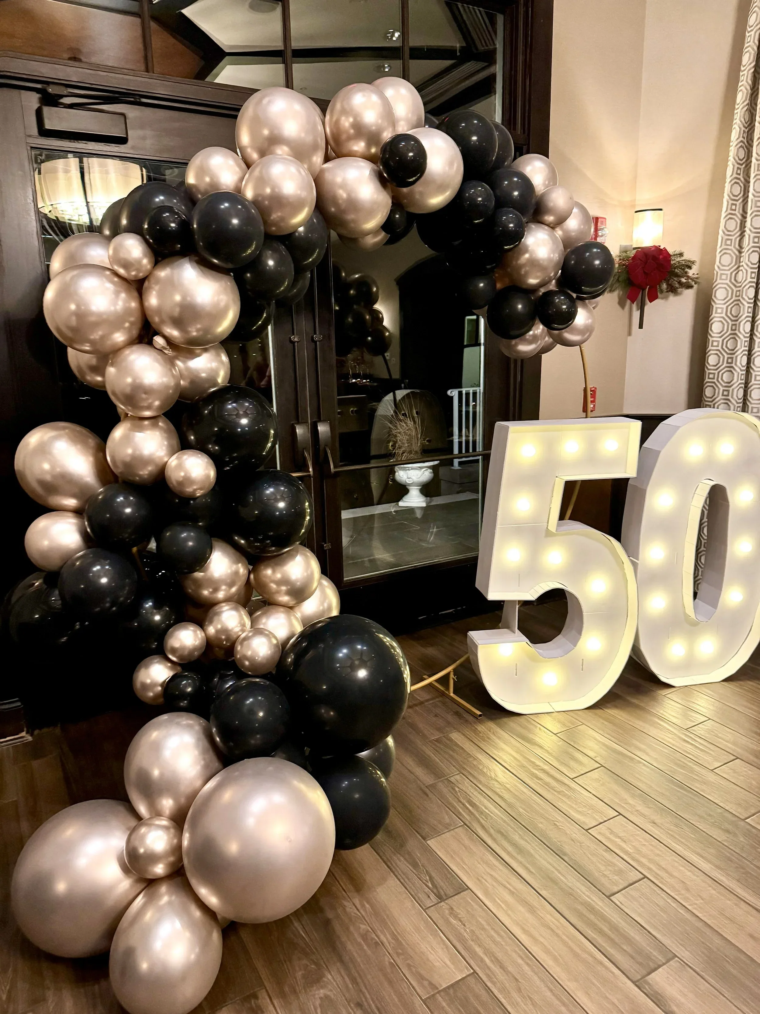 A large Balloon arch made of black, gold, and pearl-colored balloons is displayed in front of a mirror. There are also illuminated letter numbers 3 and 6 lit with bulbs, a Christmas decoration with a red bow, and a lit lamp in the background.
