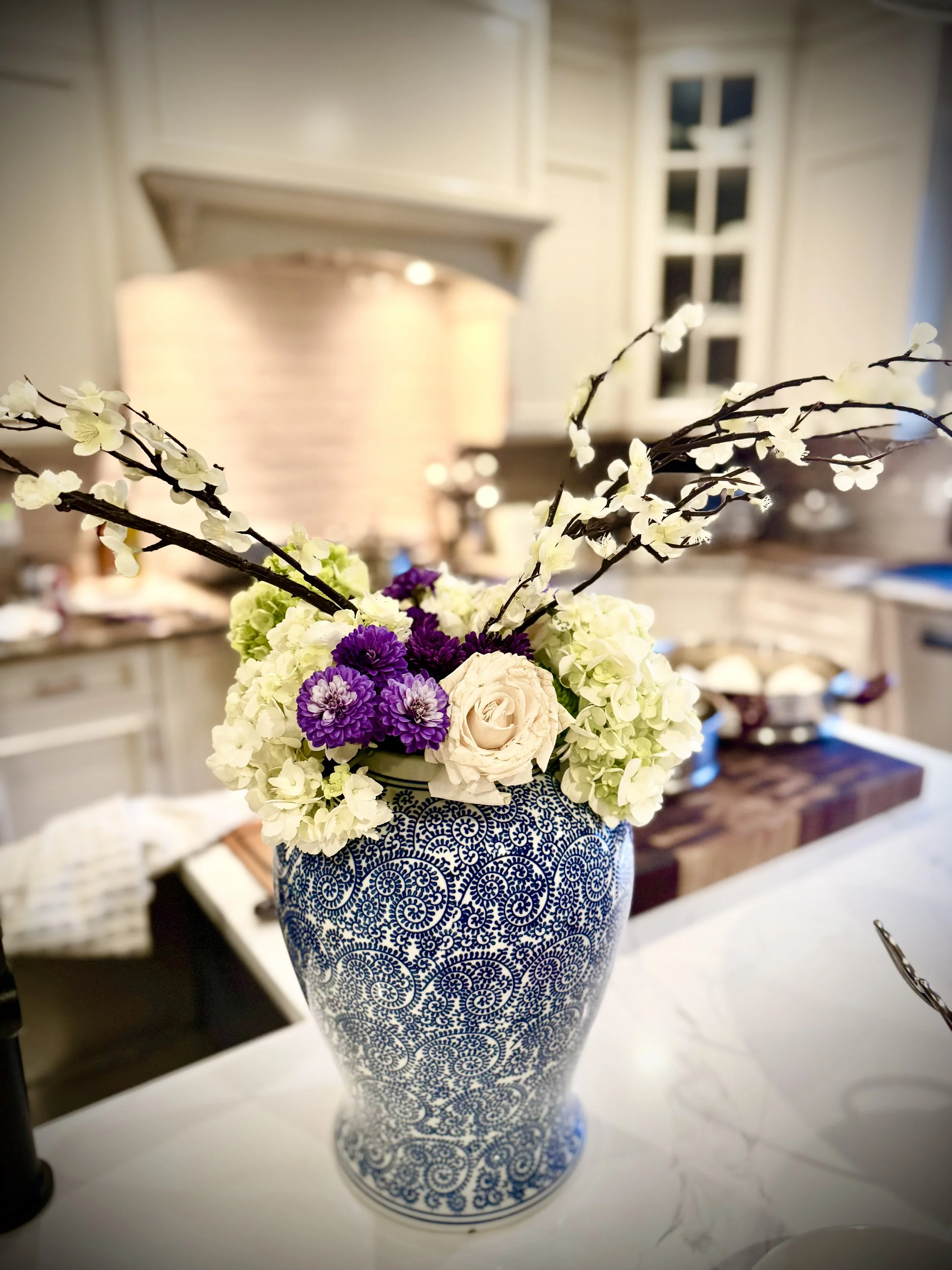 A floral arrangement in a blue and white patterned vase on a kitchen countertop with a blurred background of kitchen cabinets and a window.