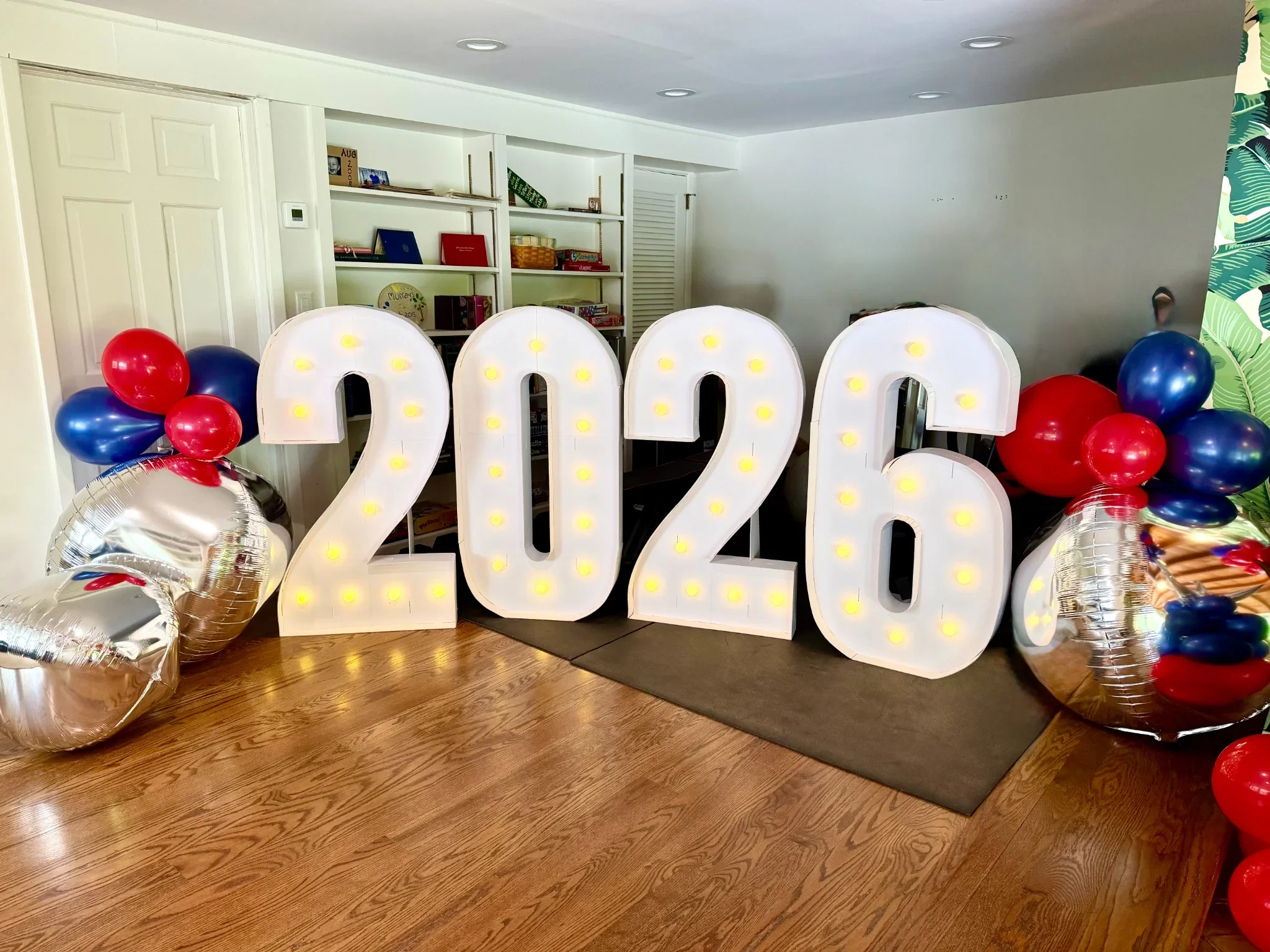 Decorative graduation display with large illuminated numbers '2023', surrounded by red, blue, and silver balloons, in a room with white walls, a bookshelf, and wood flooring.