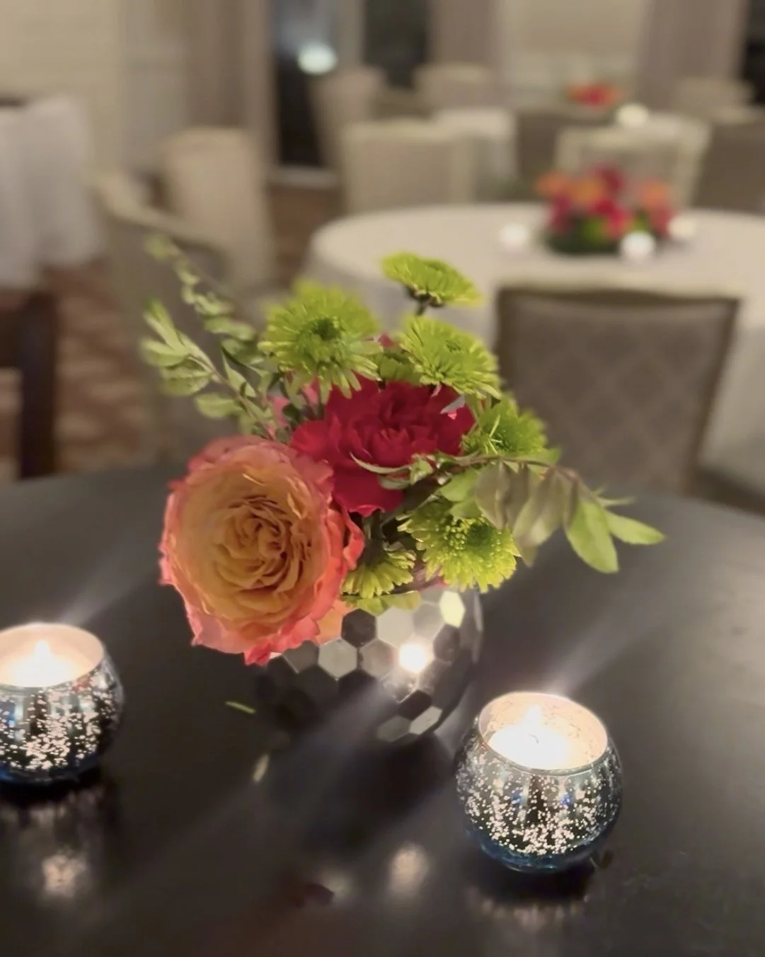 A floral centerpiece with pink, orange, and green flowers on a black table, flanked by two small lit candles in decorative holders, in a dimly lit room with round tables and chairs.