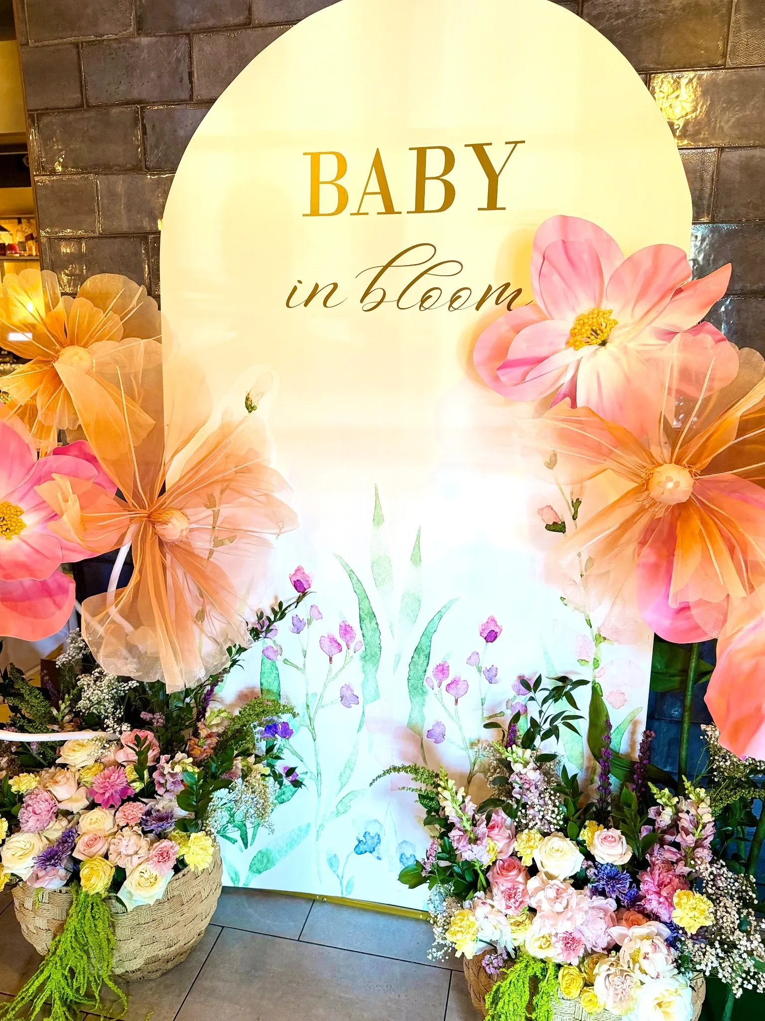 A floral display with a large arrangement of pink, purple, yellow, and white flowers in wicker baskets on a tiled floor. Behind is a white display board with the words 'BABY in bloom' and an illustration of pink flowers.