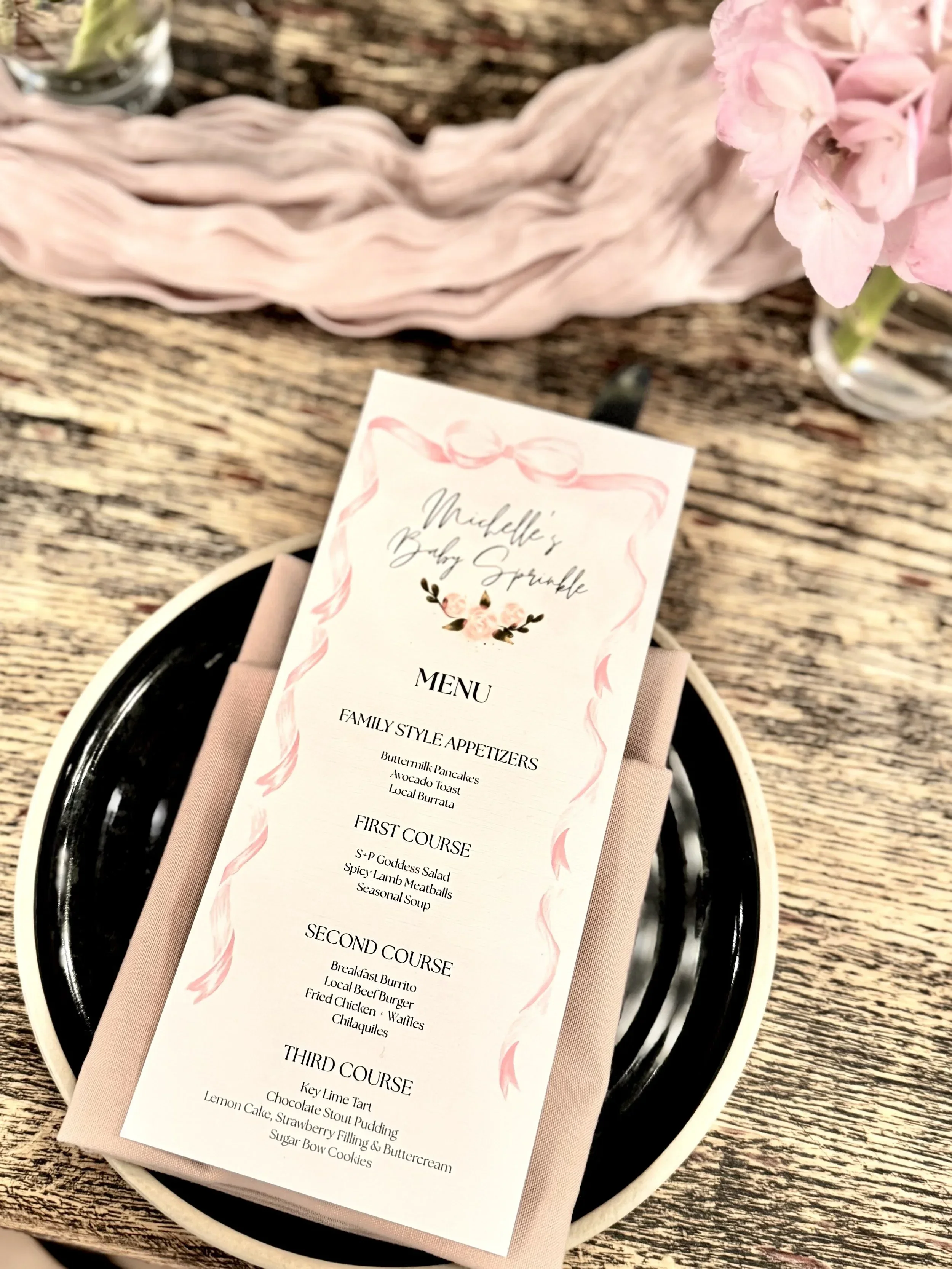 A wedding menu card resting on a pink napkin and black plate, featuring a pink watercolor border and floral design, placed on a wooden table with a pink flower and glass of water nearby.