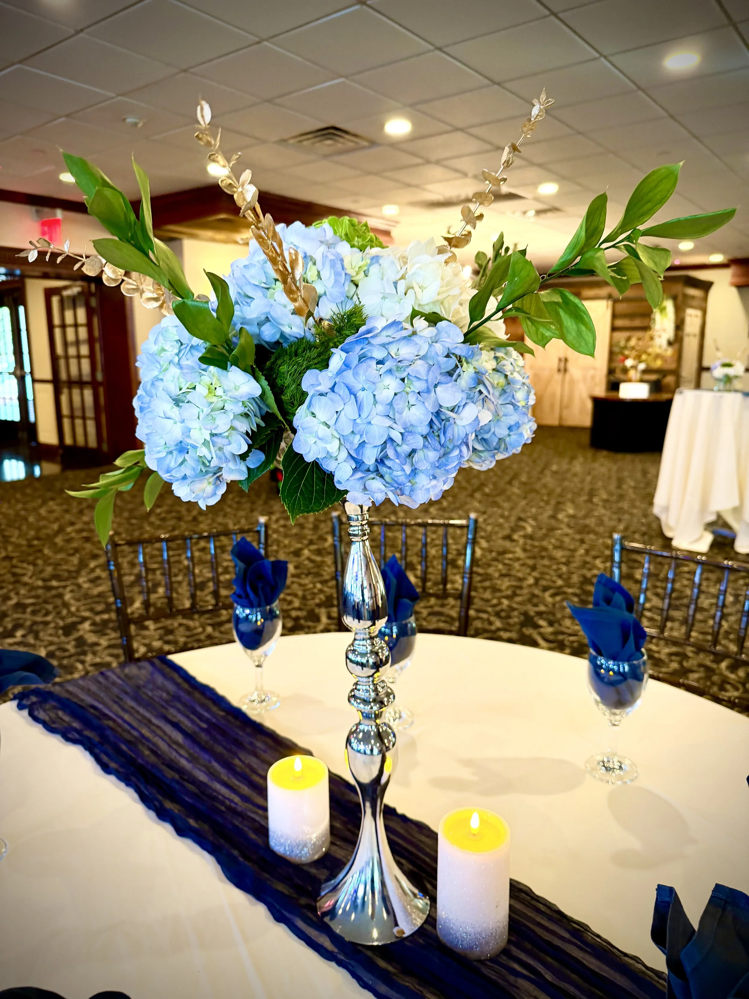 A centerpiece on a round table with blue hydrangeas, green leaves, gold and glass accents, surrounded by blue napkins in glasses, with yellow candles, set up for a formal event in an indoor banquet hall.