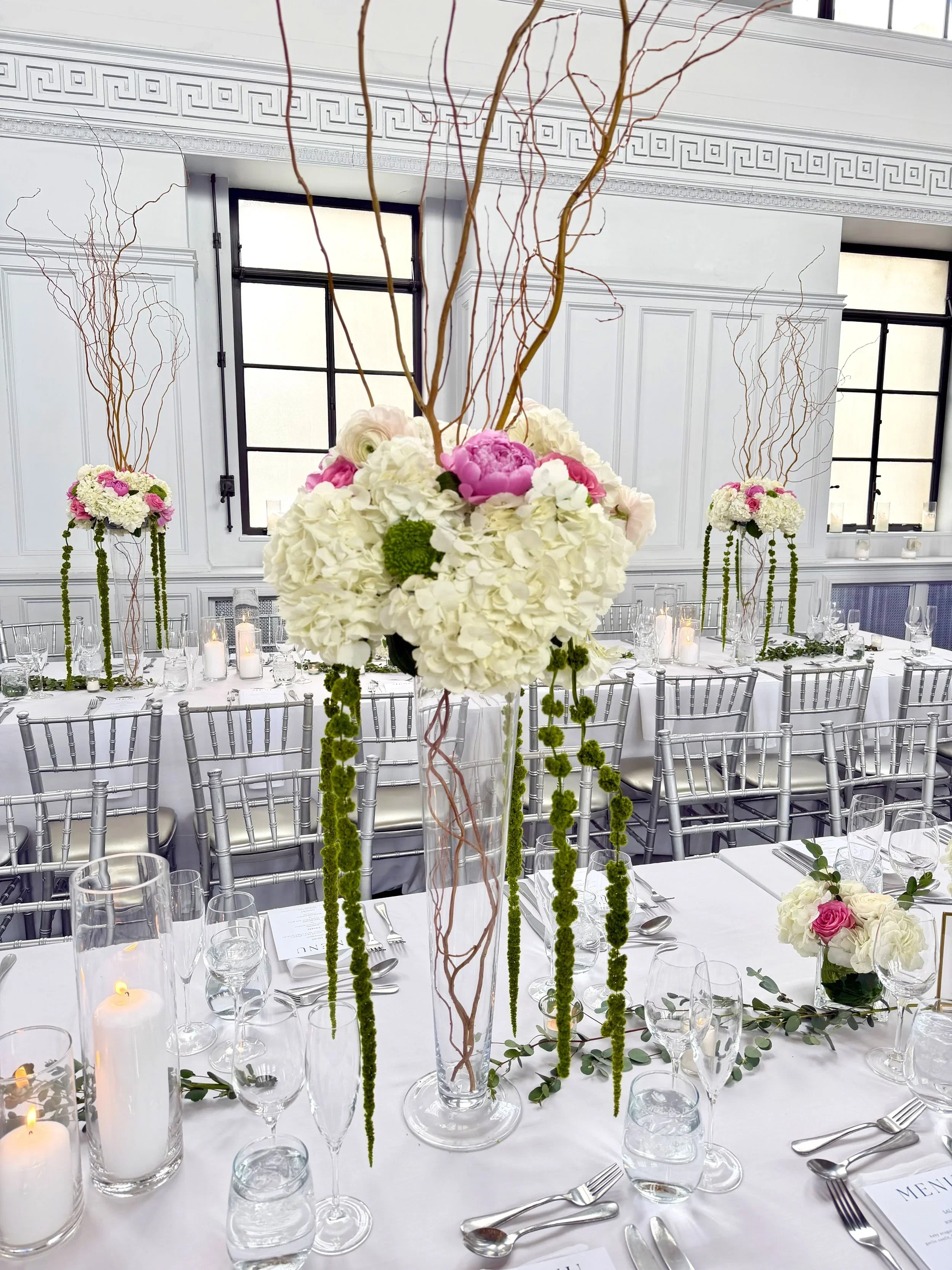 Elegant indoor wedding reception table decor featuring a tall floral centerpiece with white hydrangeas, pink roses, and decorative branches. The table is set with white tablecloths, glassware, silverware, candles, and greenery accents.