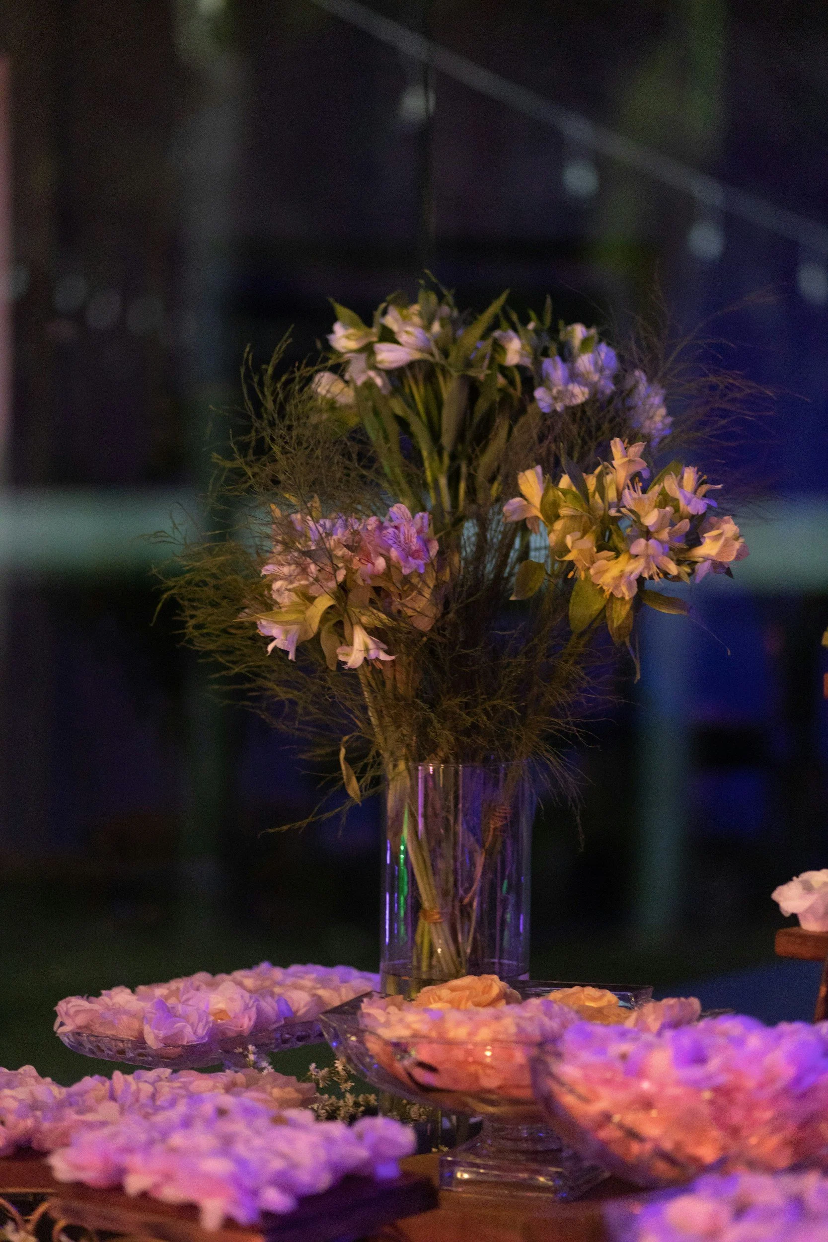 A tall glass vase holding a bouquet of pink and white flowers with green leaves, on a table surrounded by glass bowls filled with flower petals, in a dimly lit setting.