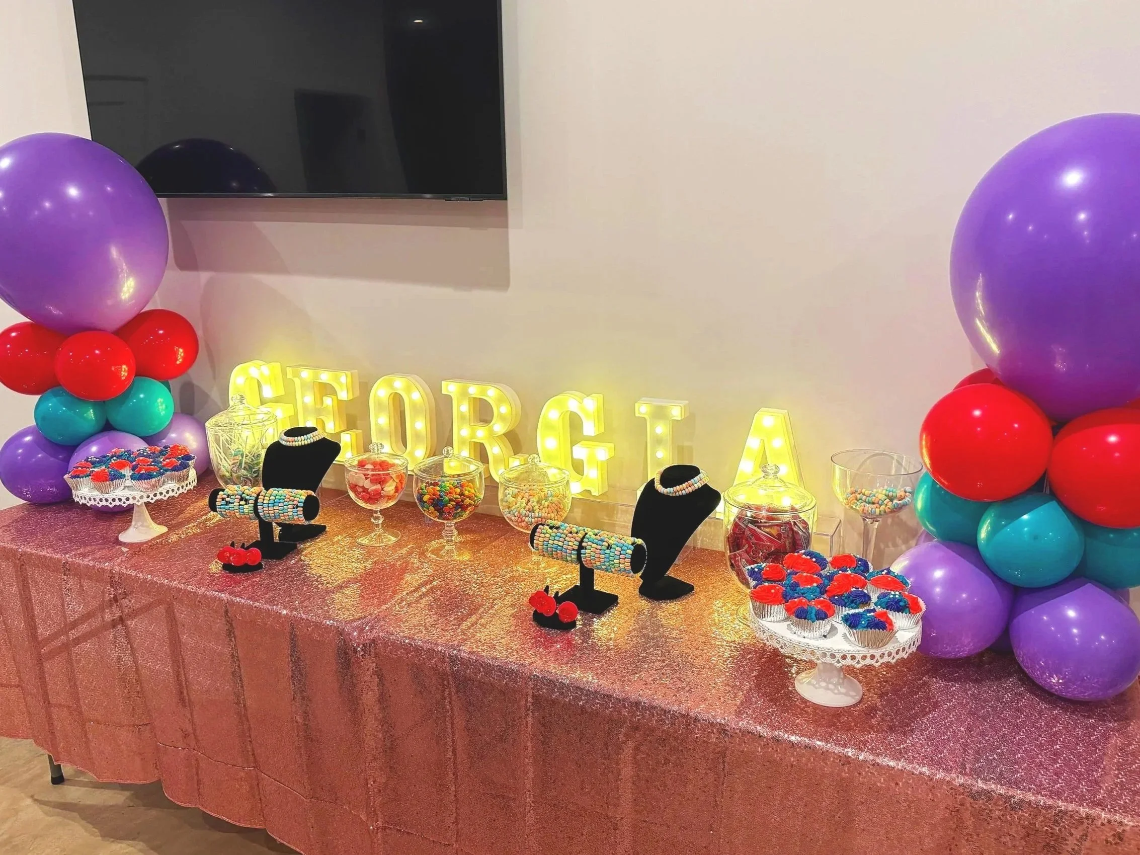 Decorated table with candy jars, cupcakes, jewelry displays, and a sign spelling 'GEORGIA' in illuminated letters, flanked by colorful balloon arrangements on each side.