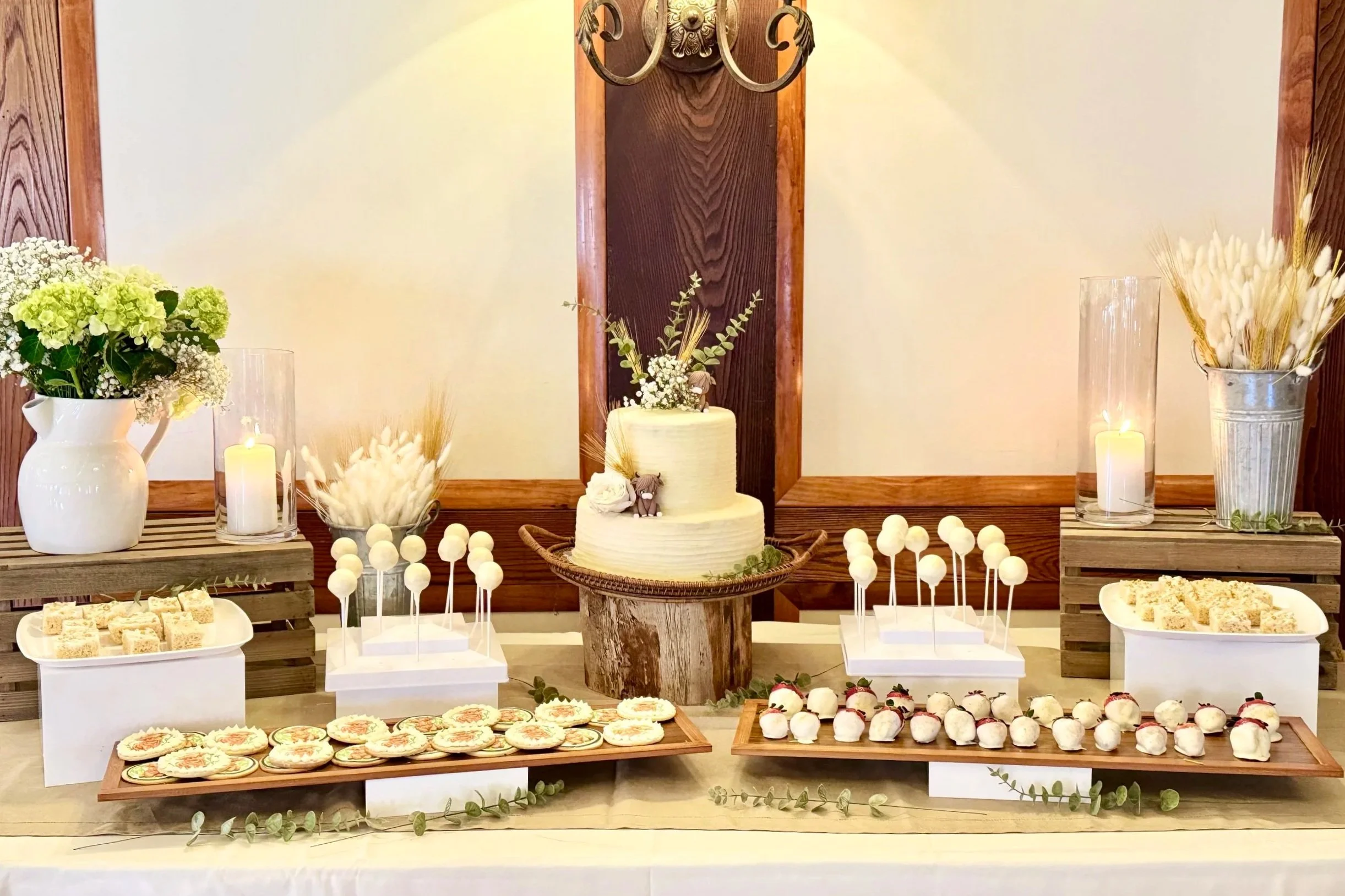 Dessert table with white cake, decorated with small flowers and an elephant figurine, surrounded by assorted cookies and treats, flanked by floral arrangements and candles in a rustic setting.