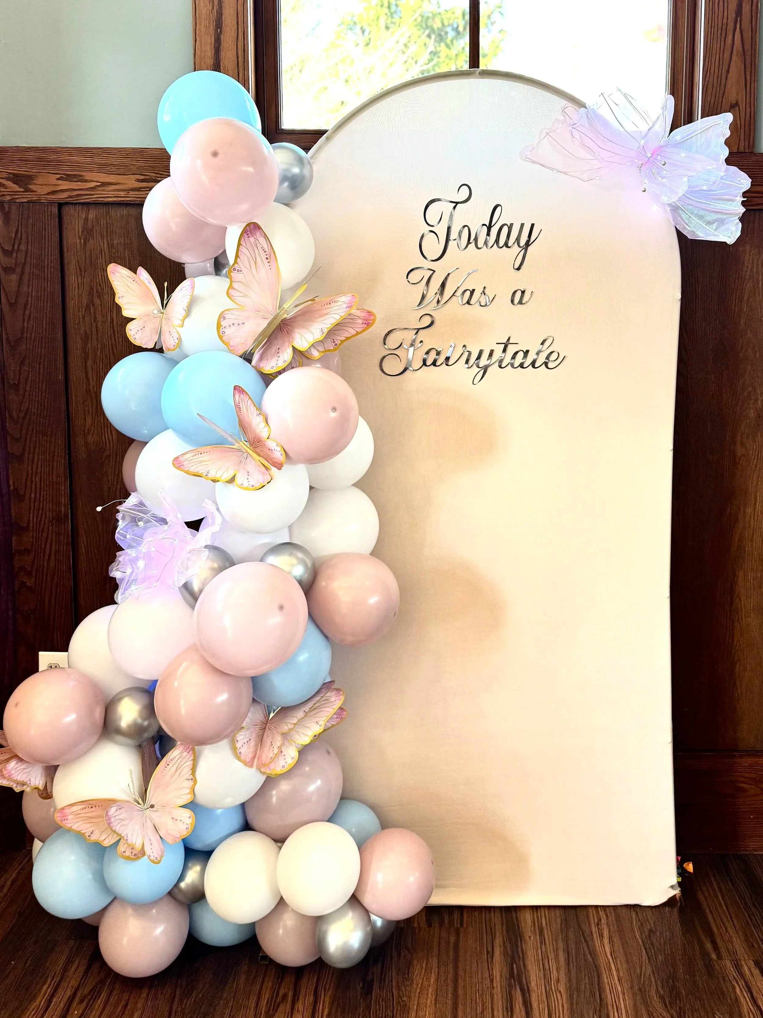 Decorative setup with pastel-colored balloons and pink butterfly cutouts on a cream backdrop that reads 'Today Was a Fairytale' in cursive, positioned in front of a wooden wall and a window.