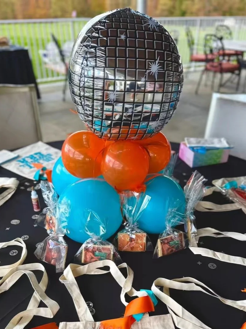 Balloon decoration featuring a large silver disco ball-shaped balloon, blue and orange balloons, with party favors and decorations on a black table.