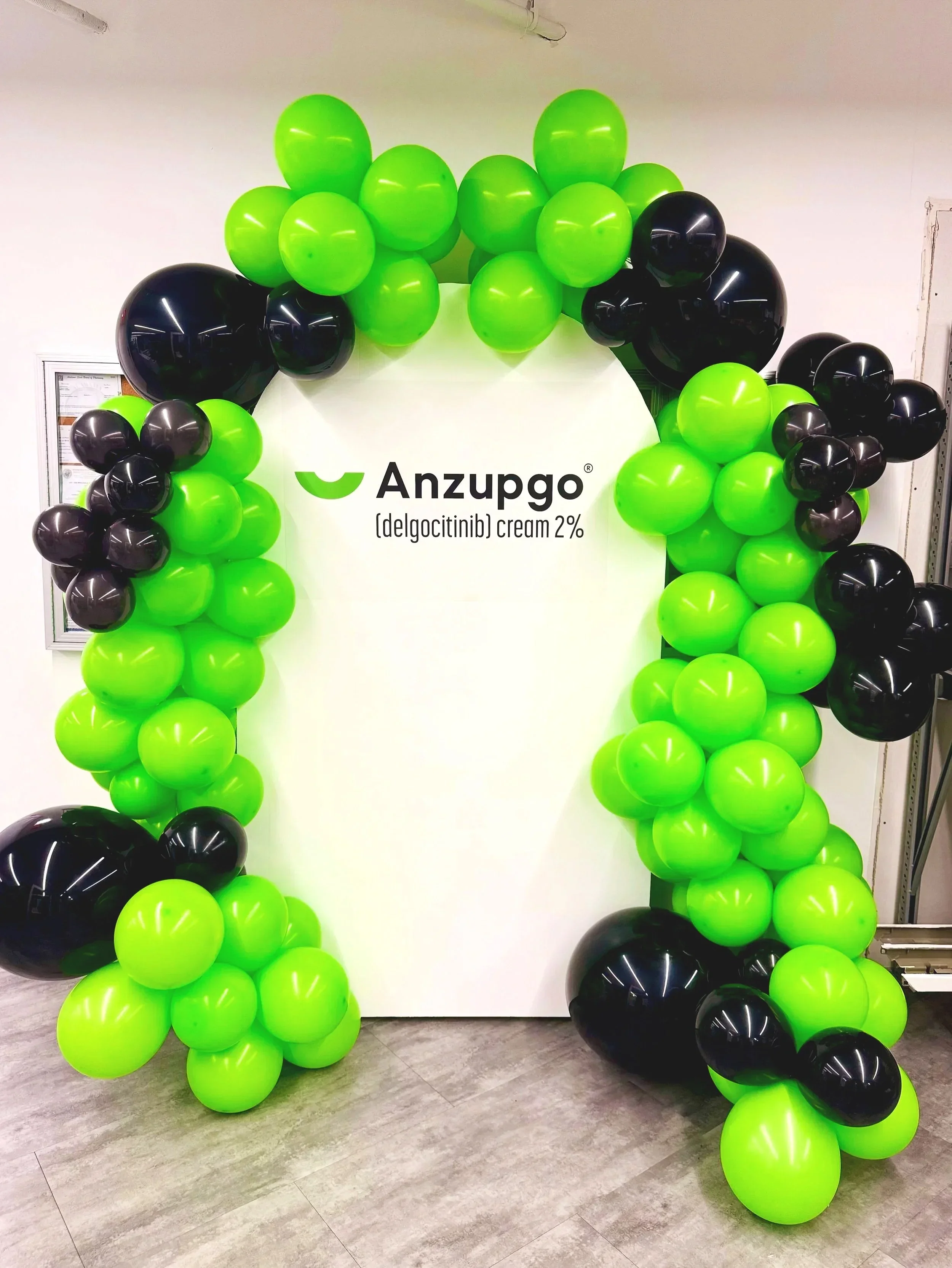 Decorative arch made of green and black balloons with the logo and text for Anzupgo and delgocitinib 2% on a white backdrop.