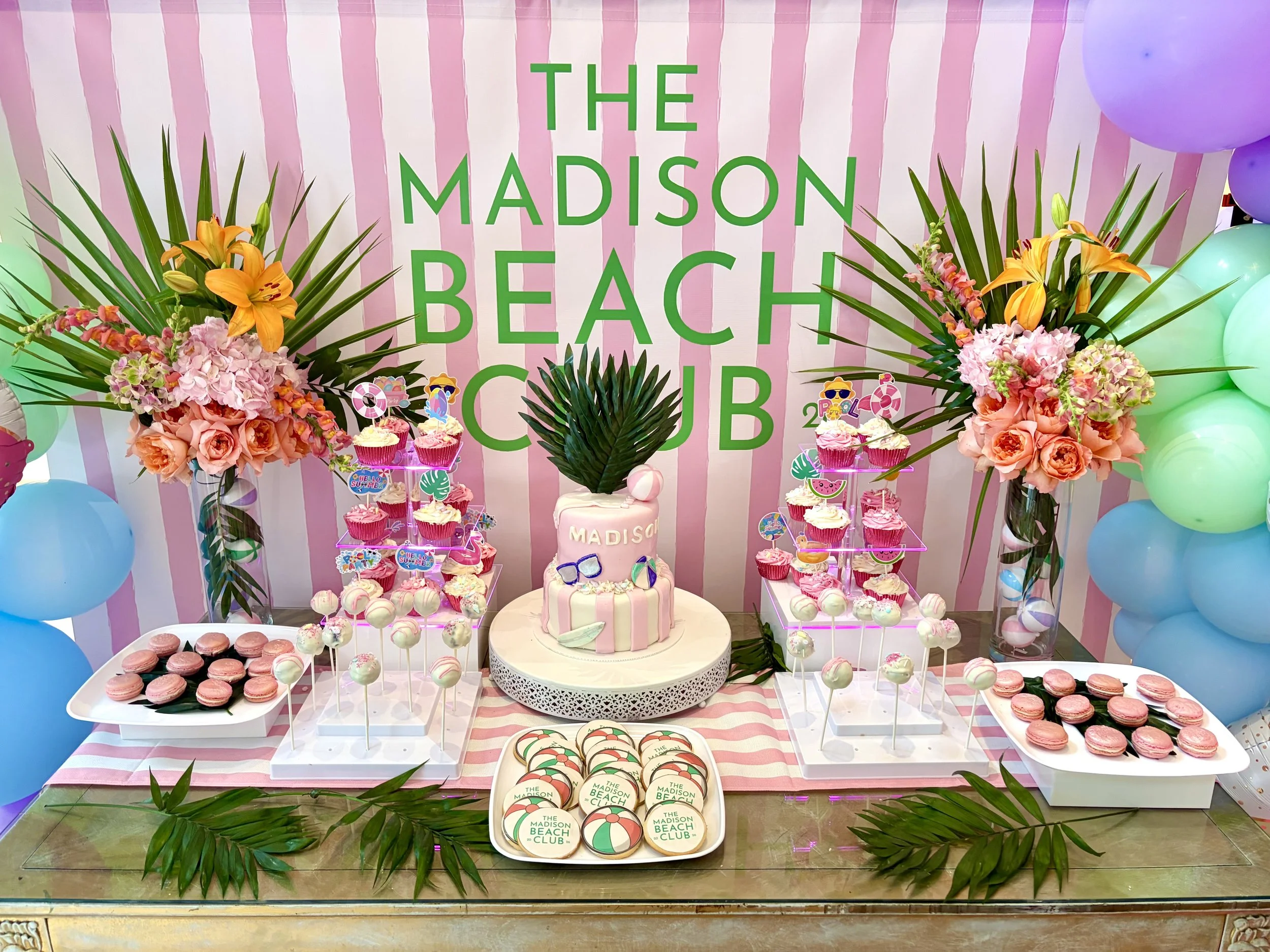 Decorative dessert table with pink and green theme, featuring a cake with 'Madison' written on it, surrounded by cupcakes, macarons, and cookies, adorned with tropical flowers and palm leaves, against a backdrop with pink stripes and green lettering 