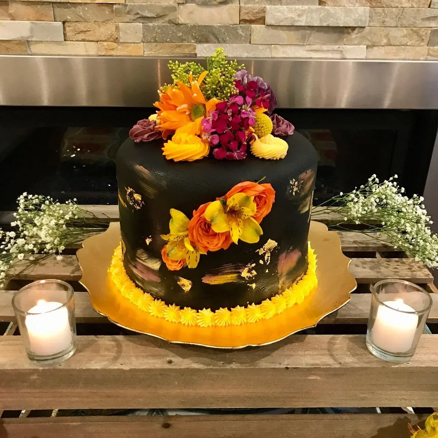 A two-tiered black wedding cake decorated with orange, yellow, pink, and purple flowers, placed on a gold cake board with yellow frosting at the base. The cake is surrounded by small white flowers and two lit candles in glass holders on a rustic wood