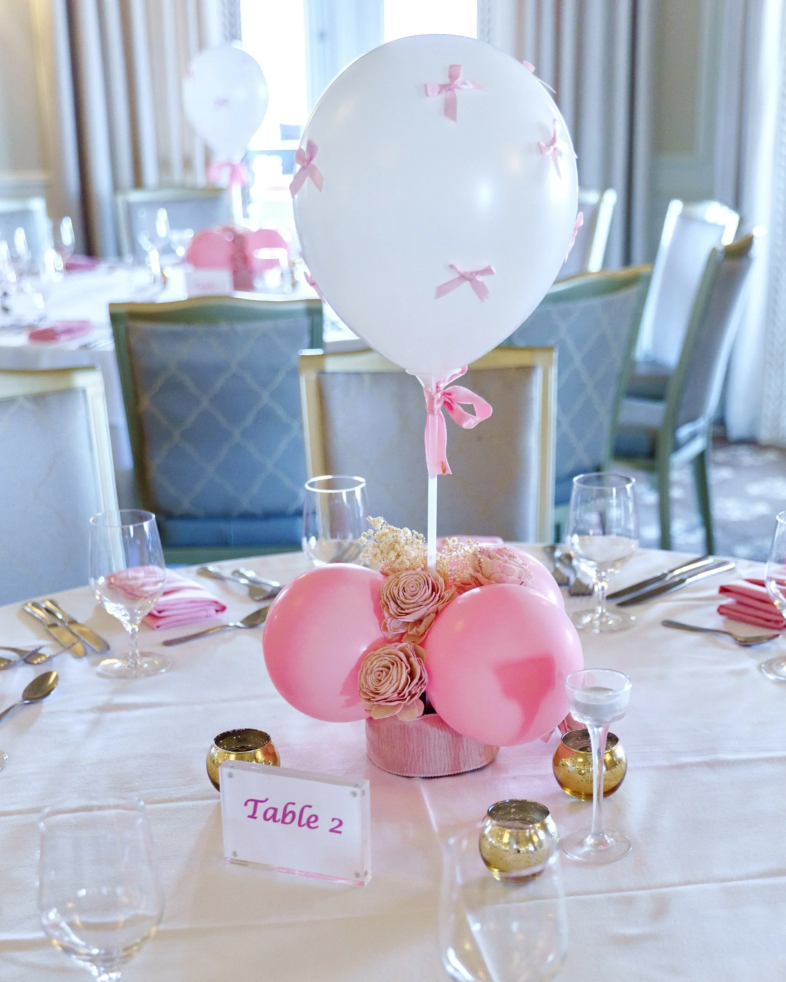 A decorated banquet table with pink balloons, pink roses, and gold candle holders, set in a bright room with chairs and tableware.