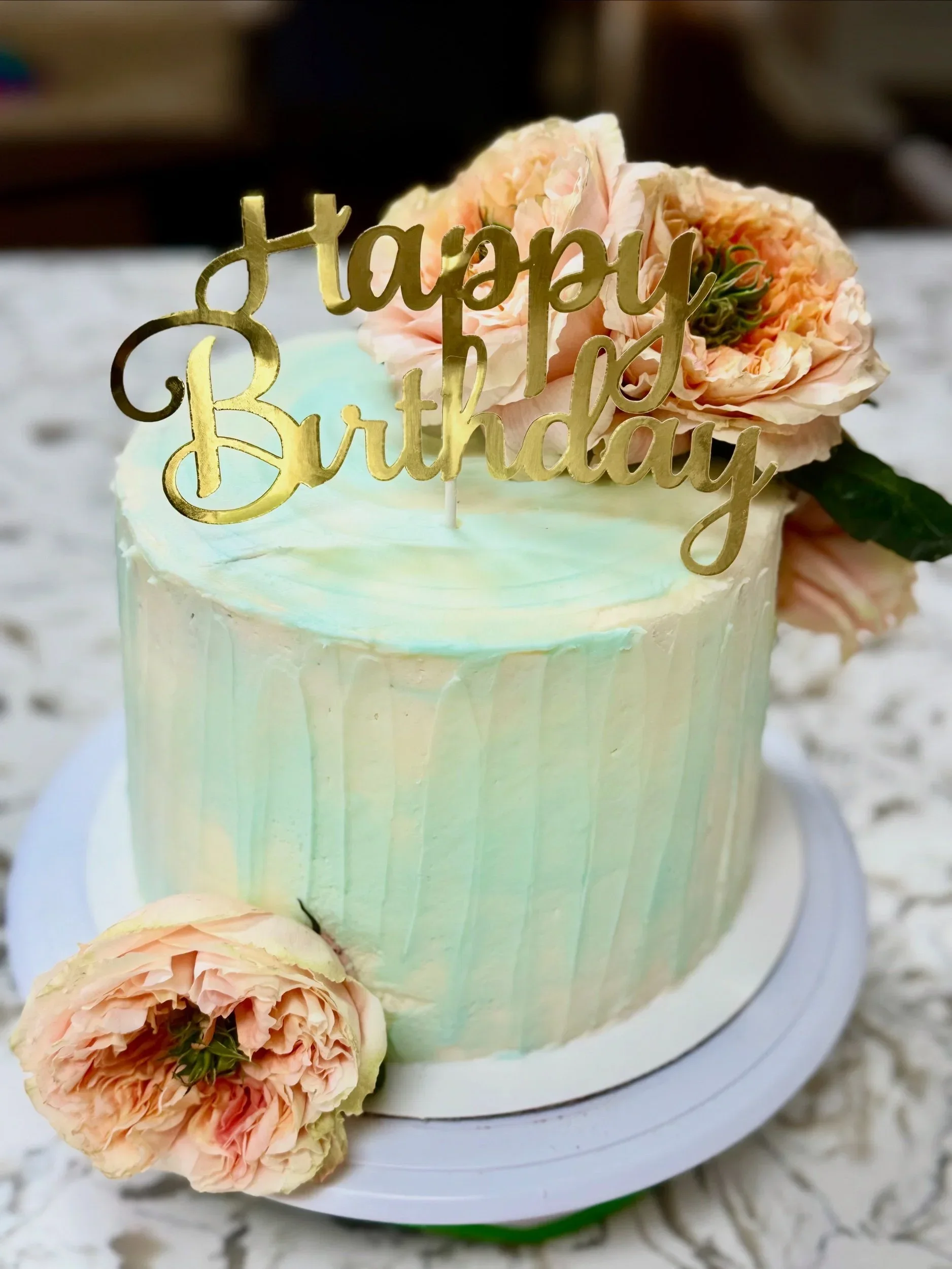 A birthday cake with a pastel green ombre frosting, decorated with pink and peach roses and a gold 'Happy Birthday' cake topper.