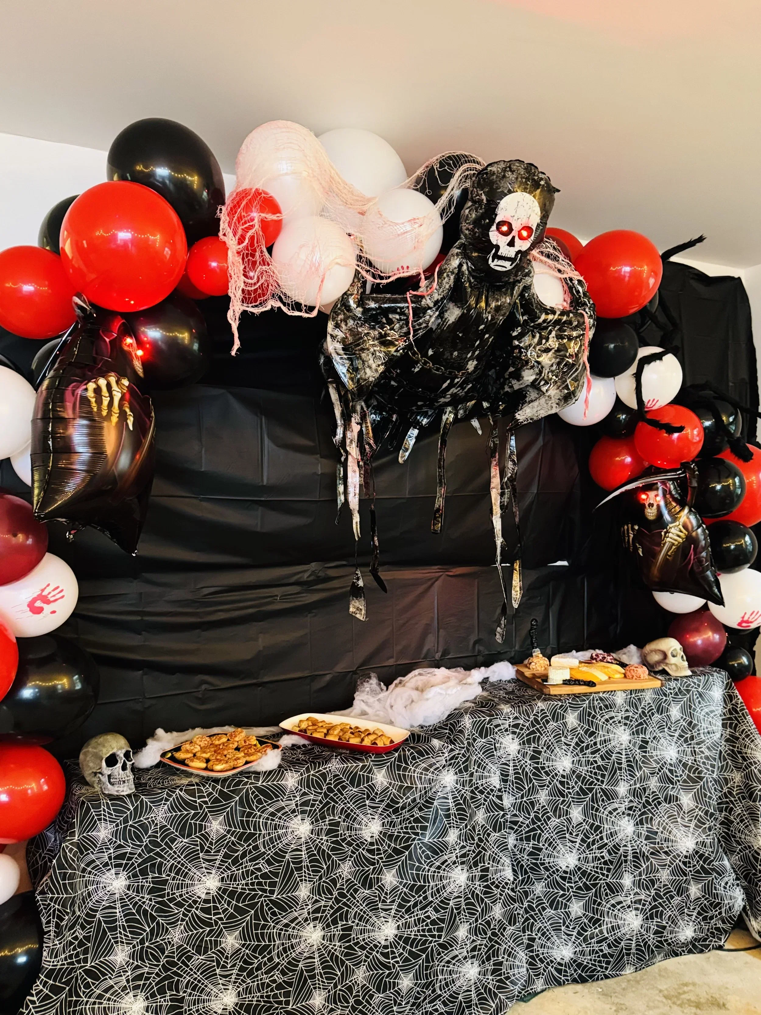 A Halloween themed party table decorated with a black and white spider web tablecloth, balloons in black, red, and white, a large spooky skeleton figure with glowing red eyes, and creepy decorations such as skulls, fake cobwebs, and chains. There are