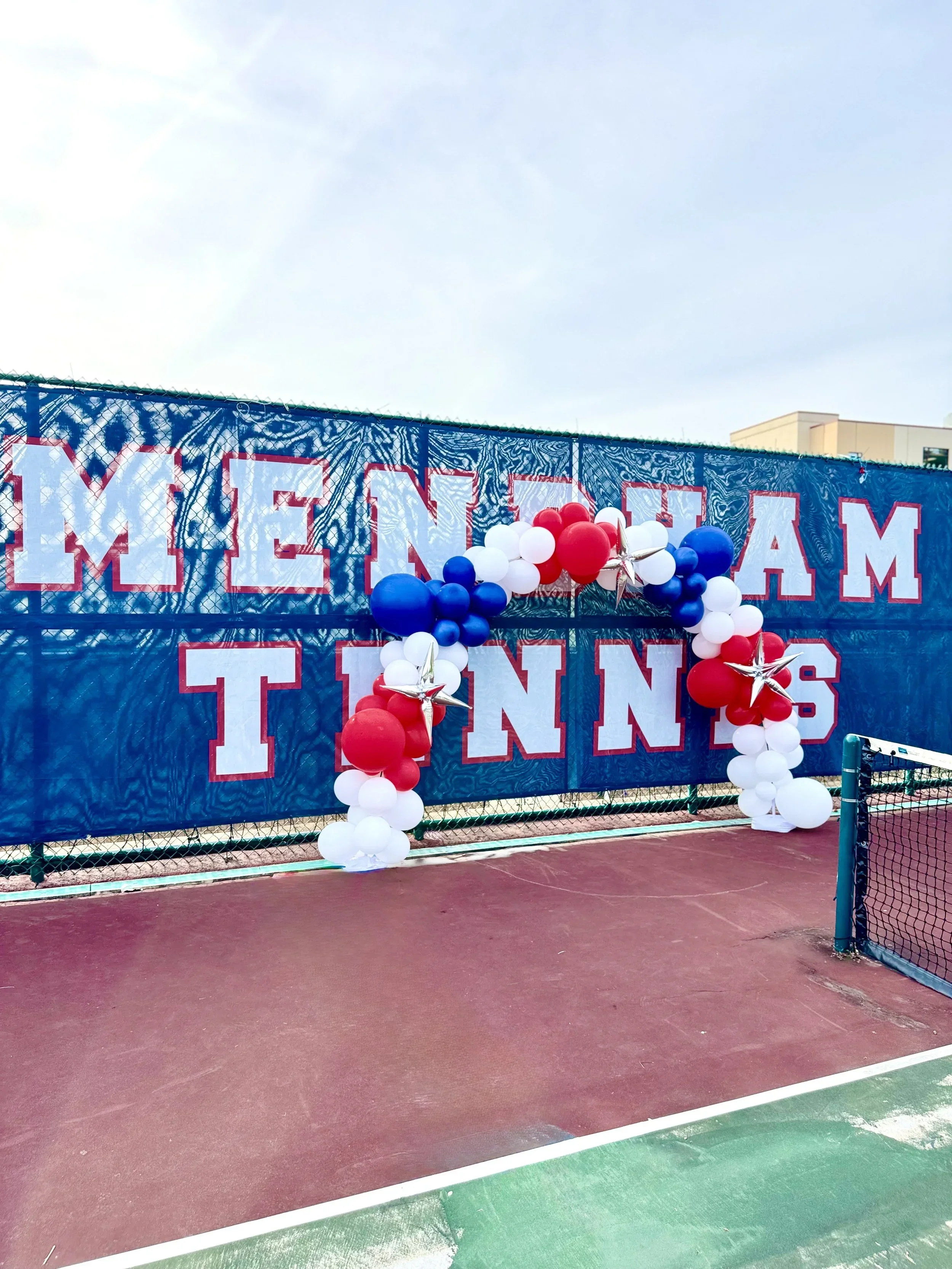 Decorated fence with red, white, and blue balloons and stars over a tennis court, with some buildings and a cloudy sky in the background.