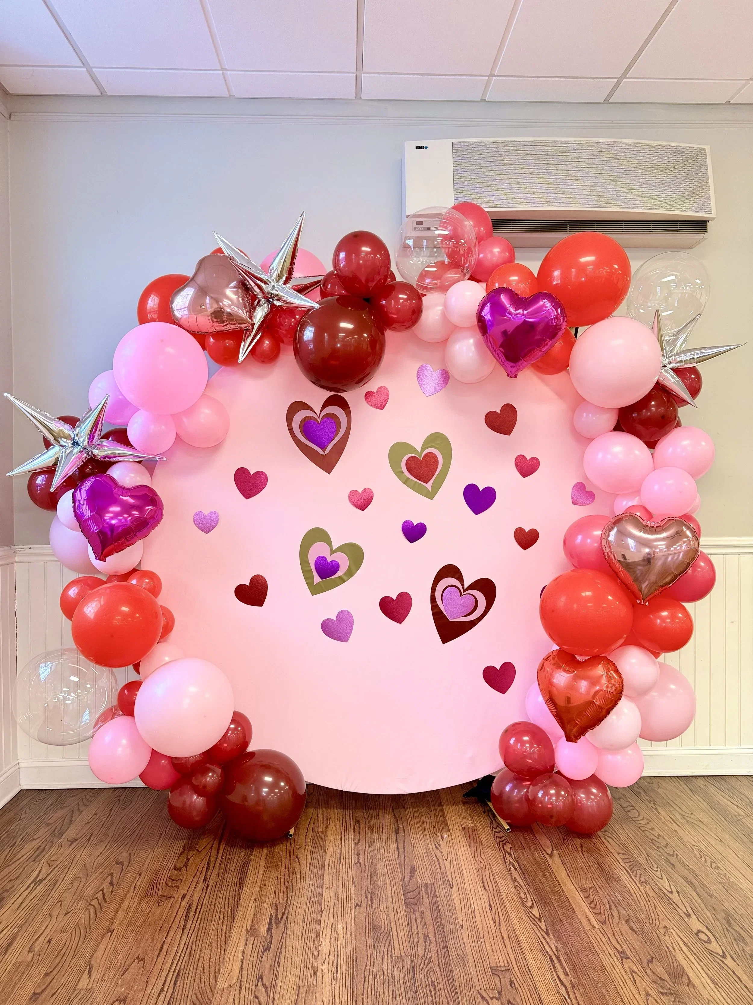 A decorated pink backdrop with red, pink, and purple balloons arranged around the edges, some shaped like hearts and stars. Heart-shaped paper cutouts are attached to the backdrop, creating a festive atmosphere.