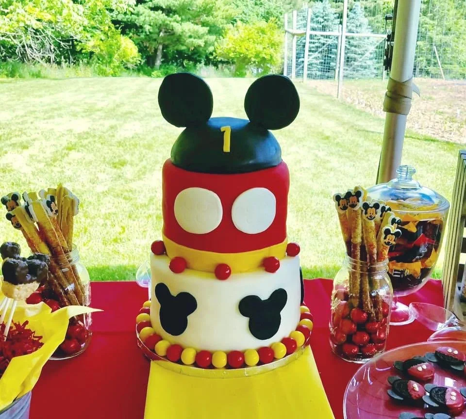 A cake decorated with Mickey Mouse theme, featuring three tiers with Mickey Mouse ears on top, red, white, yellow, and black colors, surrounded by Mickey Mouse-themed candies and snacks on a red tablecloth at an outdoor party.