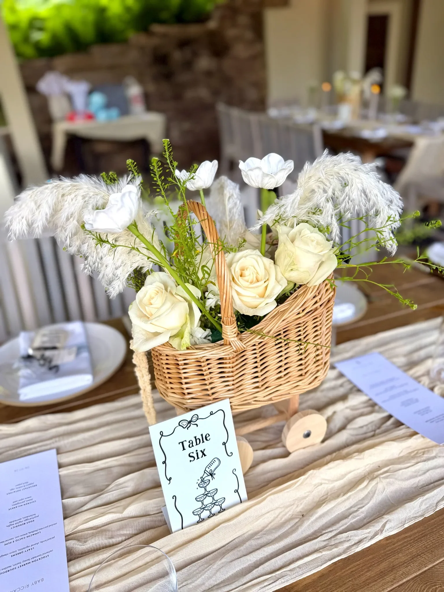 A wicker basket filled with white roses, white pampas grass, and other greenery, placed on a table with a paper sign that reads 'Table Six' and a small illustration of a champagne tower, with a blurred background of dining tables set for an outdoor e