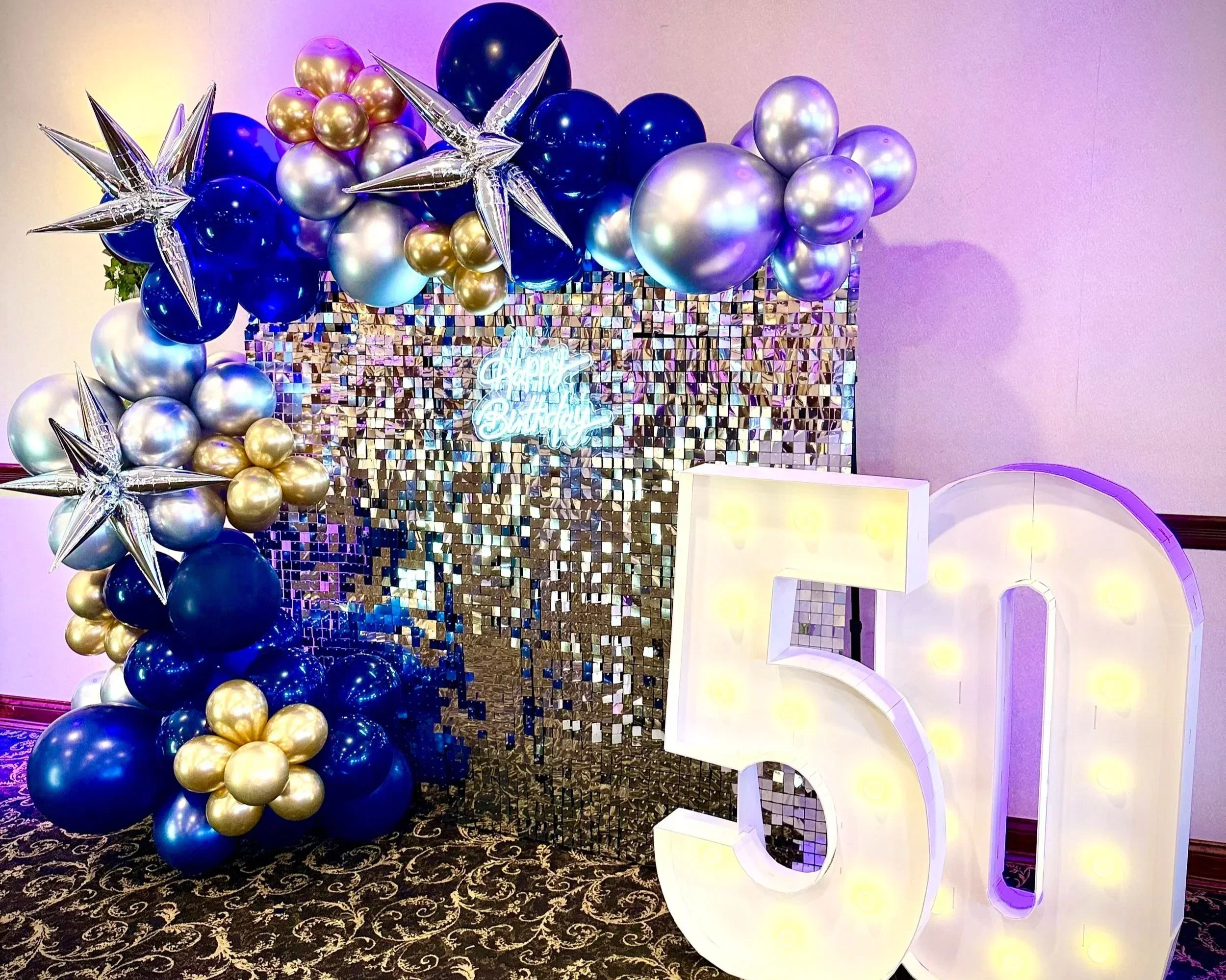 Decorative balloon arrangement with gold, silver, and navy blue balloons, silver star-shaped balloons, and a reflective backdrop with a neon sign reading 'Happy Birthday'