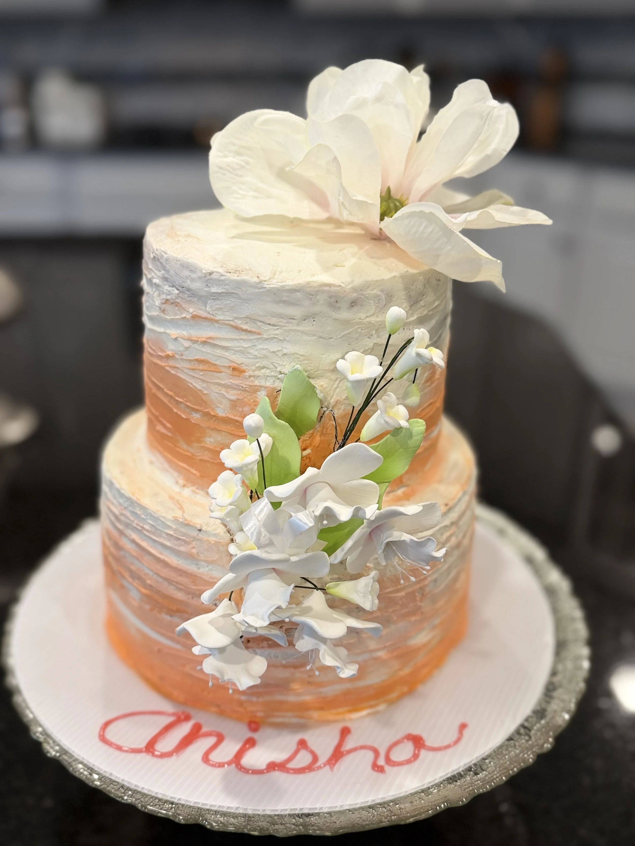 Close-up of a tiered cake decorated with white flowers and a large white flower on top, with the name 'Christo' written in red on the cake board.