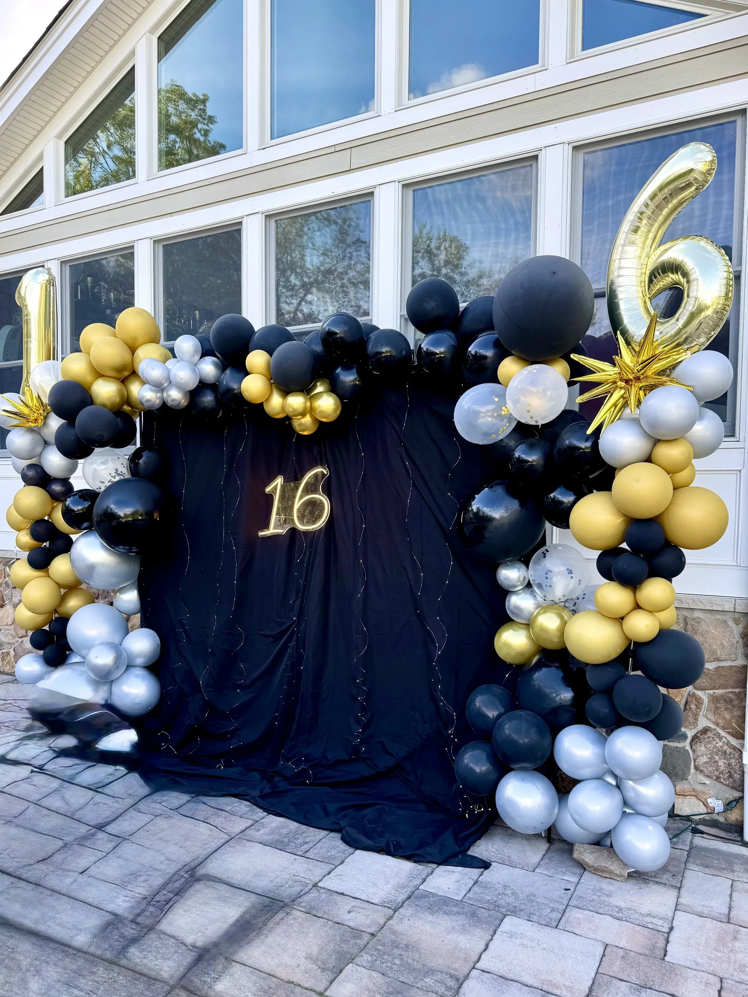 Decorative balloon arch with black, white, gold, and silver balloons in front of a house with large windows, celebrating a 16th birthday.