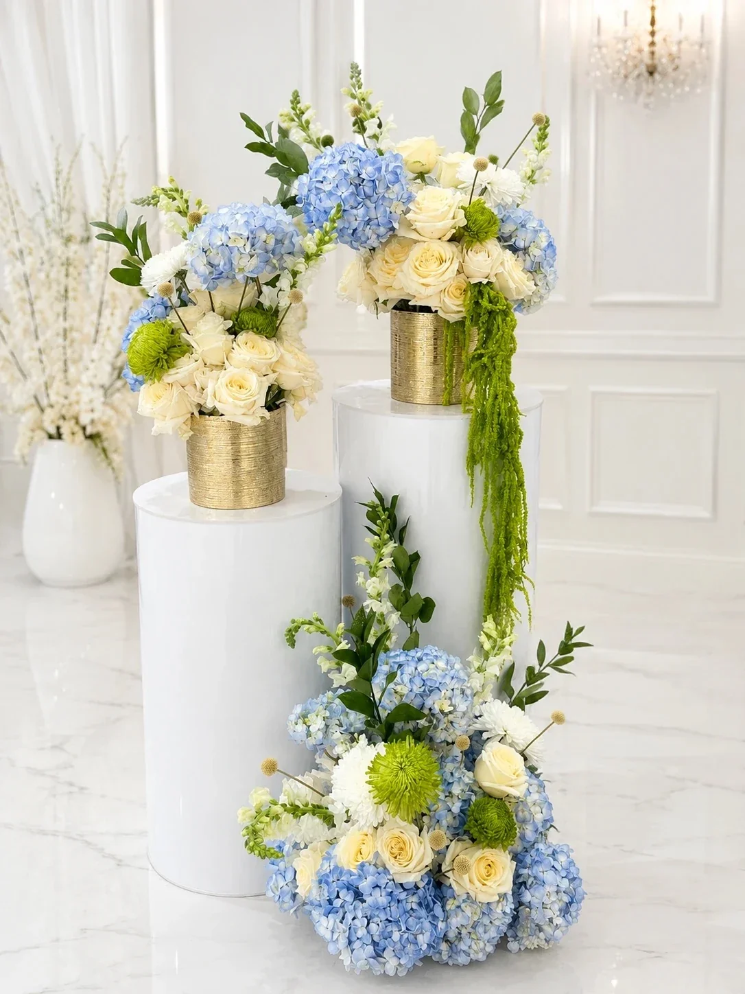 Elegant floral arrangement with blue hydrangeas, white roses, and green accents in gold vases, displayed on white cylindrical pedestals in a bright, sophisticated room.