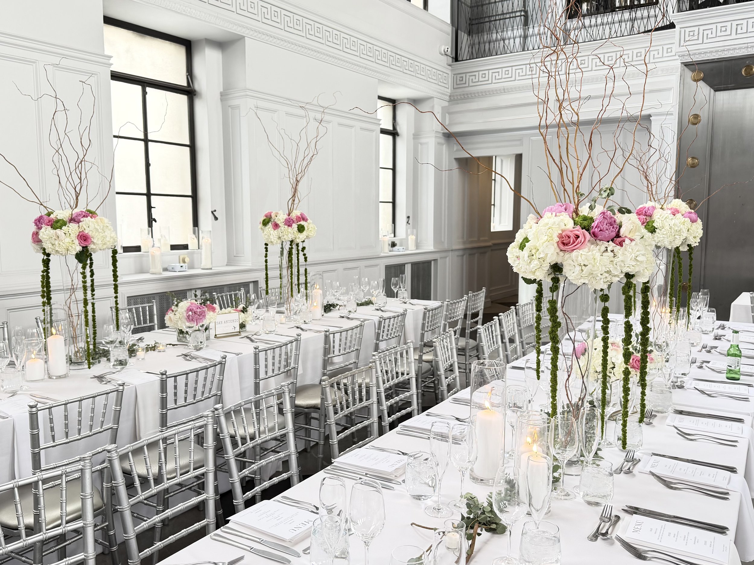 Elegant banquet setup in a bright, white-walled room with tall windows, featuring long tables with white tablecloths, silver chairs, and tall floral centerpieces with white hydrangeas, pink roses, and hanging branches. Candles and place settings are 