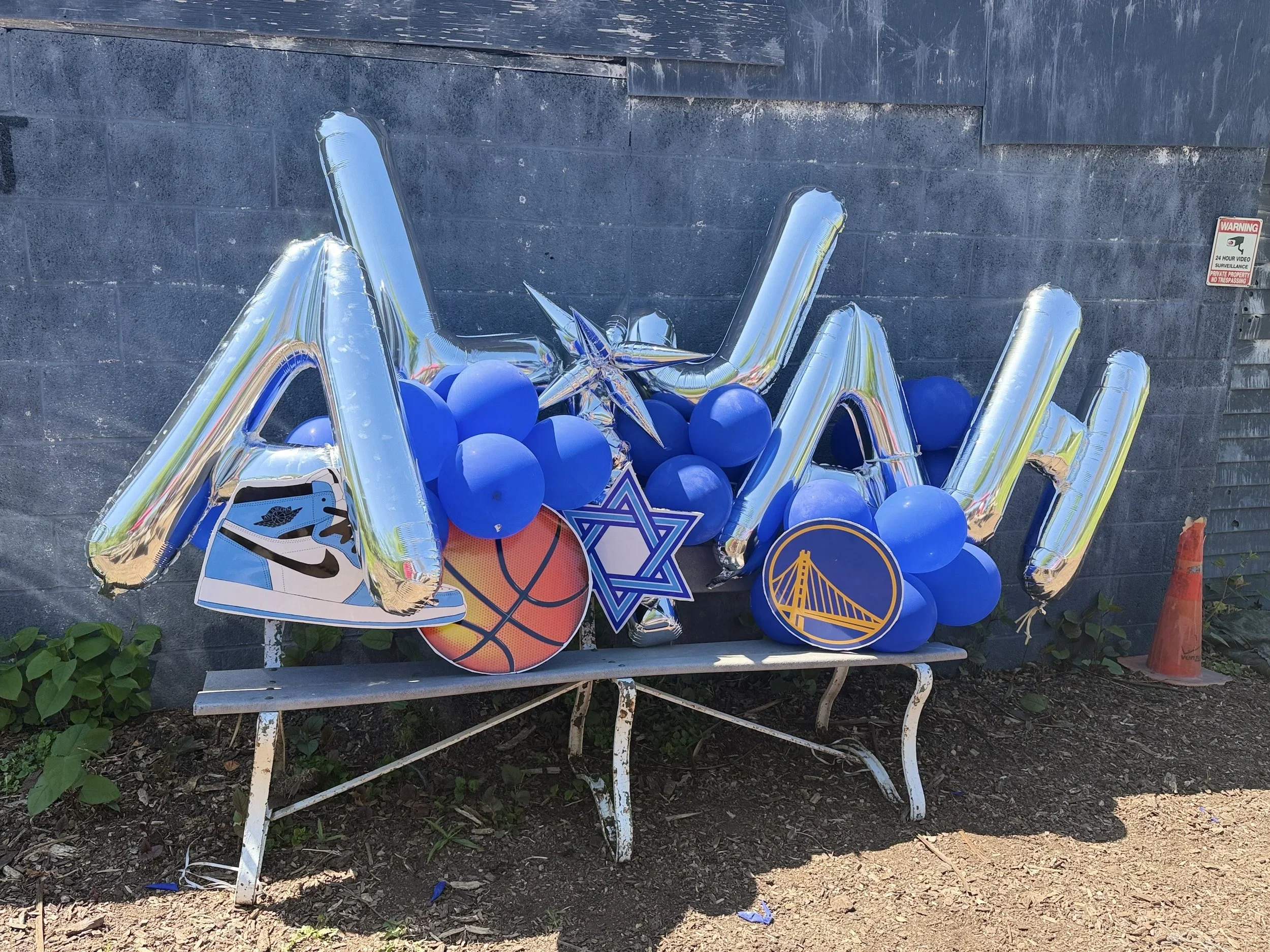 A collection of balloons, including large silver star-shaped balloons, blue round balloons, and various themed balloons, is arranged against a gray concrete wall on a white metal bench outdoors.