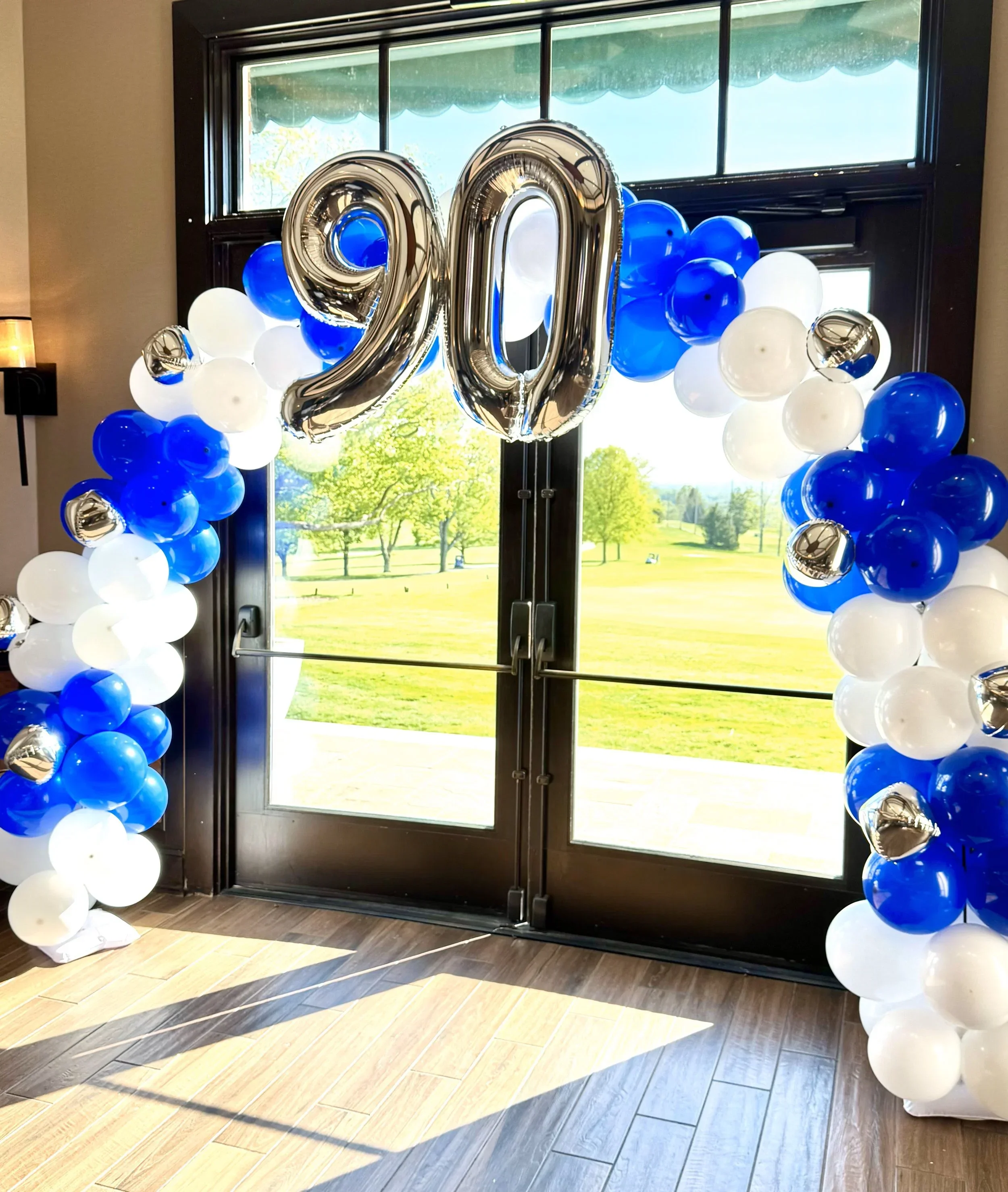 Decorative balloon arch with a large silver number 20 balloon in the center, featuring clusters of blue and white balloons on either side, set against a glass door and a bright outdoor scene.