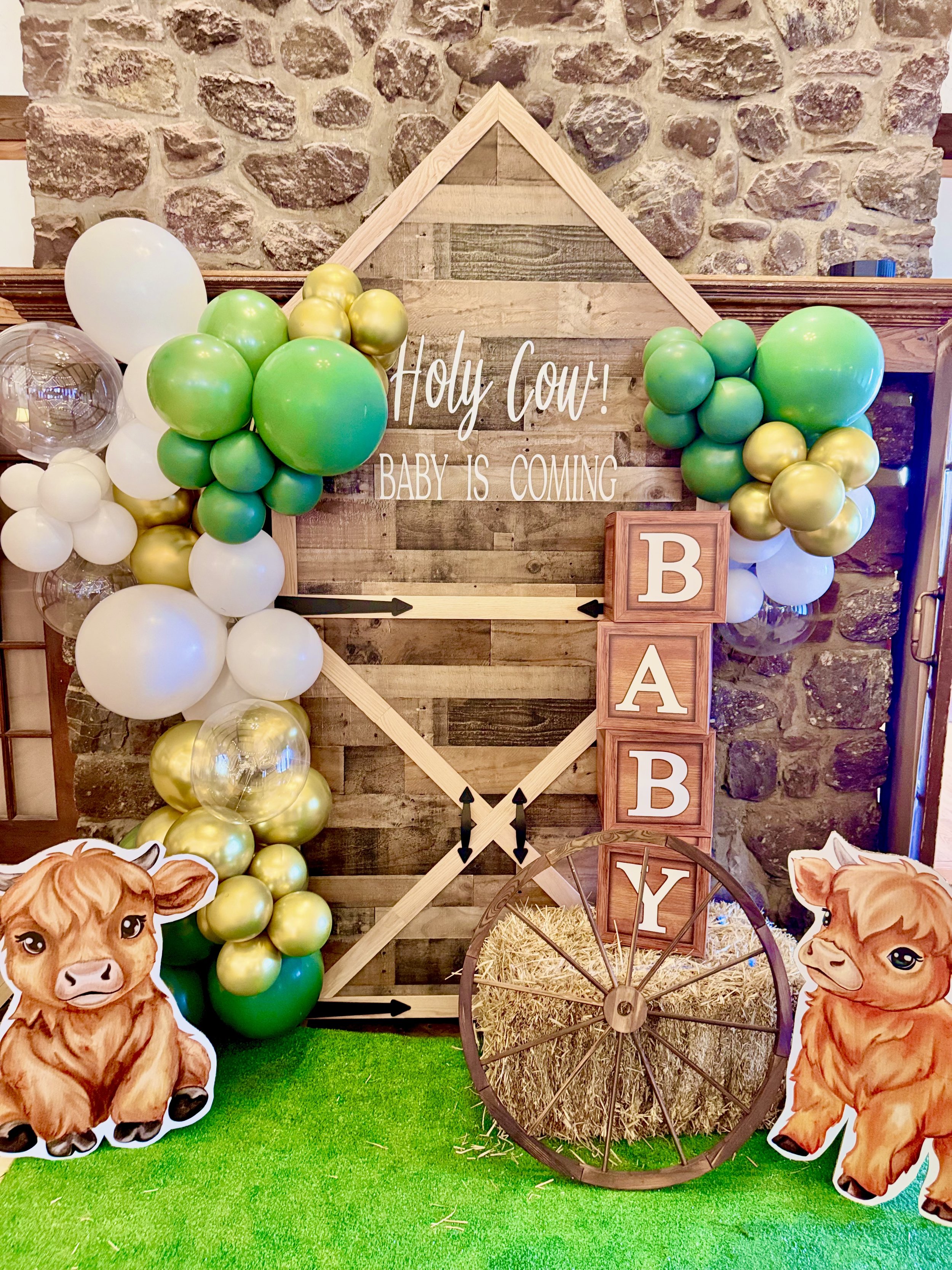 Decorative baby shower setup with wooden sign reading "Holy Cow! Baby is Coming," surrounded by green, white, and gold balloons, cartoon calf cutouts, and a wooden wagon wheel against a stone wall.