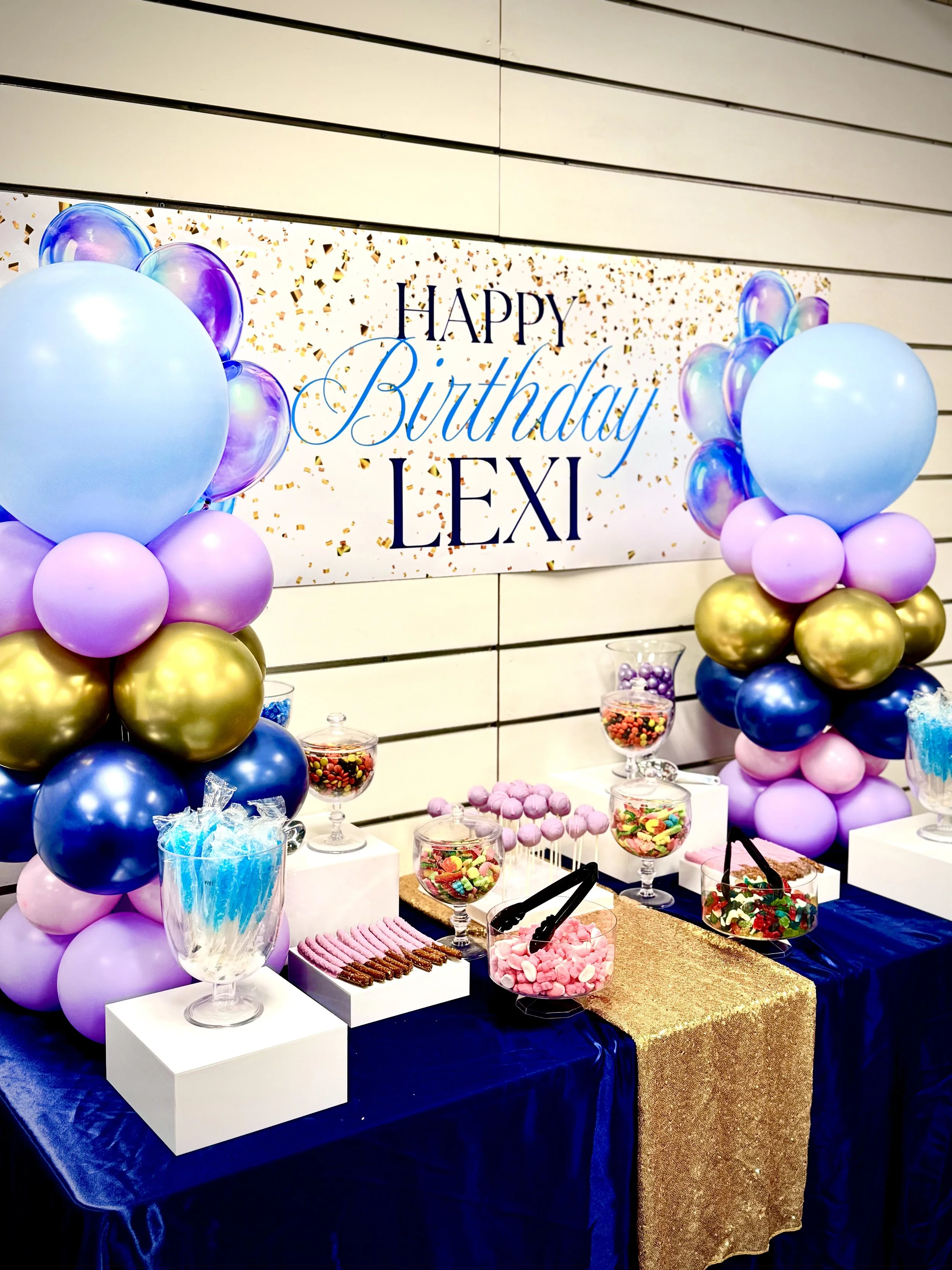 Decorated birthday party table with pink, purple, gold, and blue balloons on each side, a navy blue tablecloth, a gold sequin overlay, and a variety of candies and treats displayed on white stands and glass jars. A large sign in the background reads 