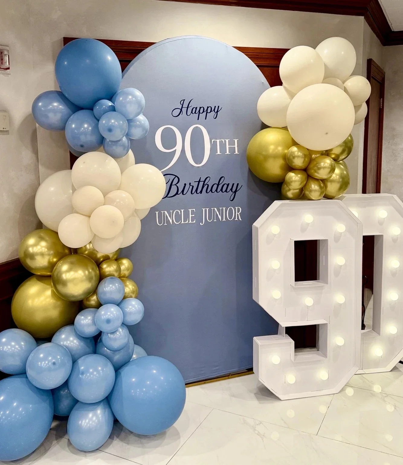 Celebration decor with a blue backdrop, clusters of blue, white, and gold balloons, a large illuminated letter 'Q', and a sign that reads 'Happy 90th Birthday Uncle Junior.'