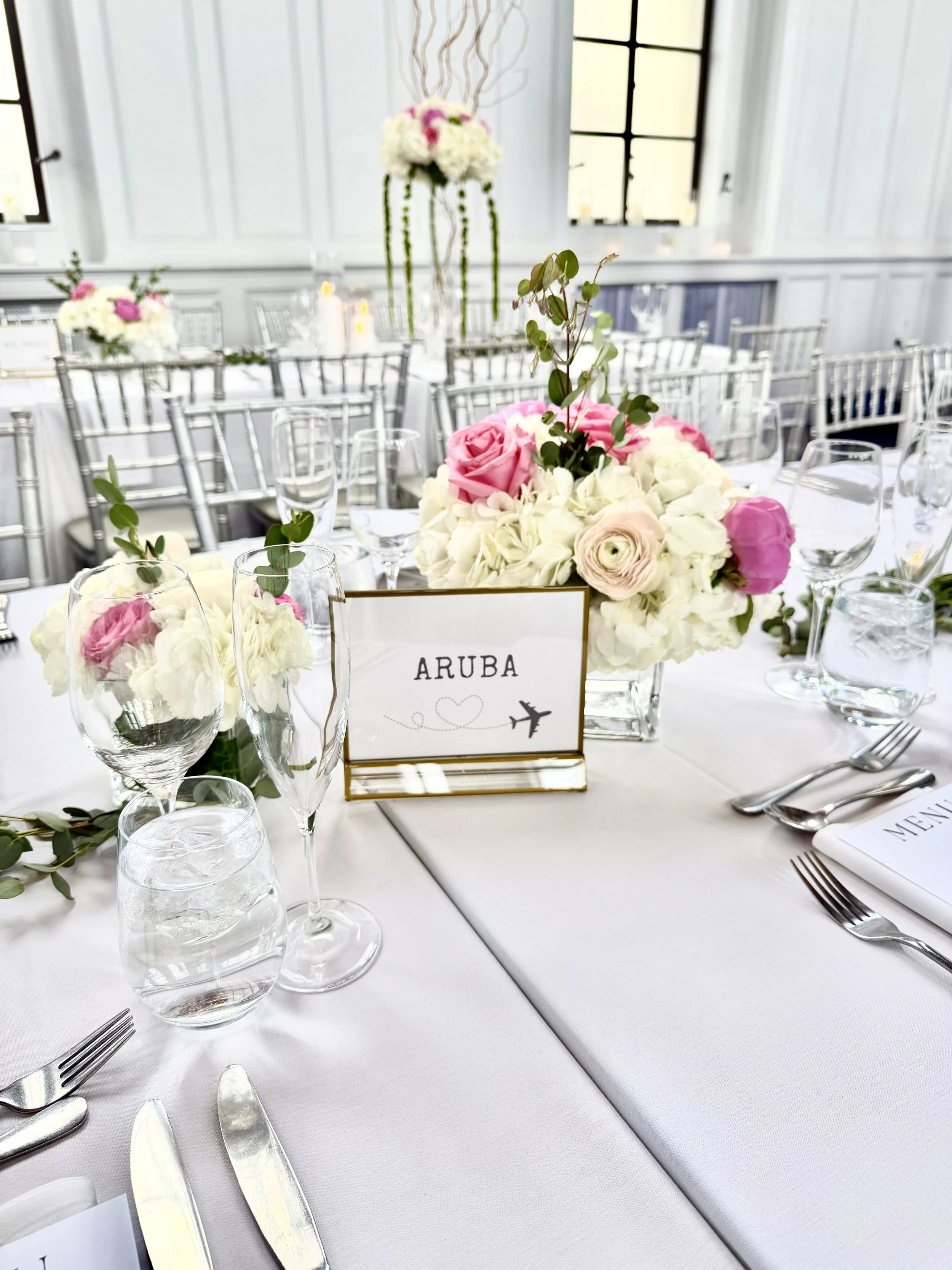 Elegant wedding table setting with white tablecloth, floral centerpiece with pink and white roses, surrounded by wine glasses, water glasses, silverware, and a sign that says "ARUBA."