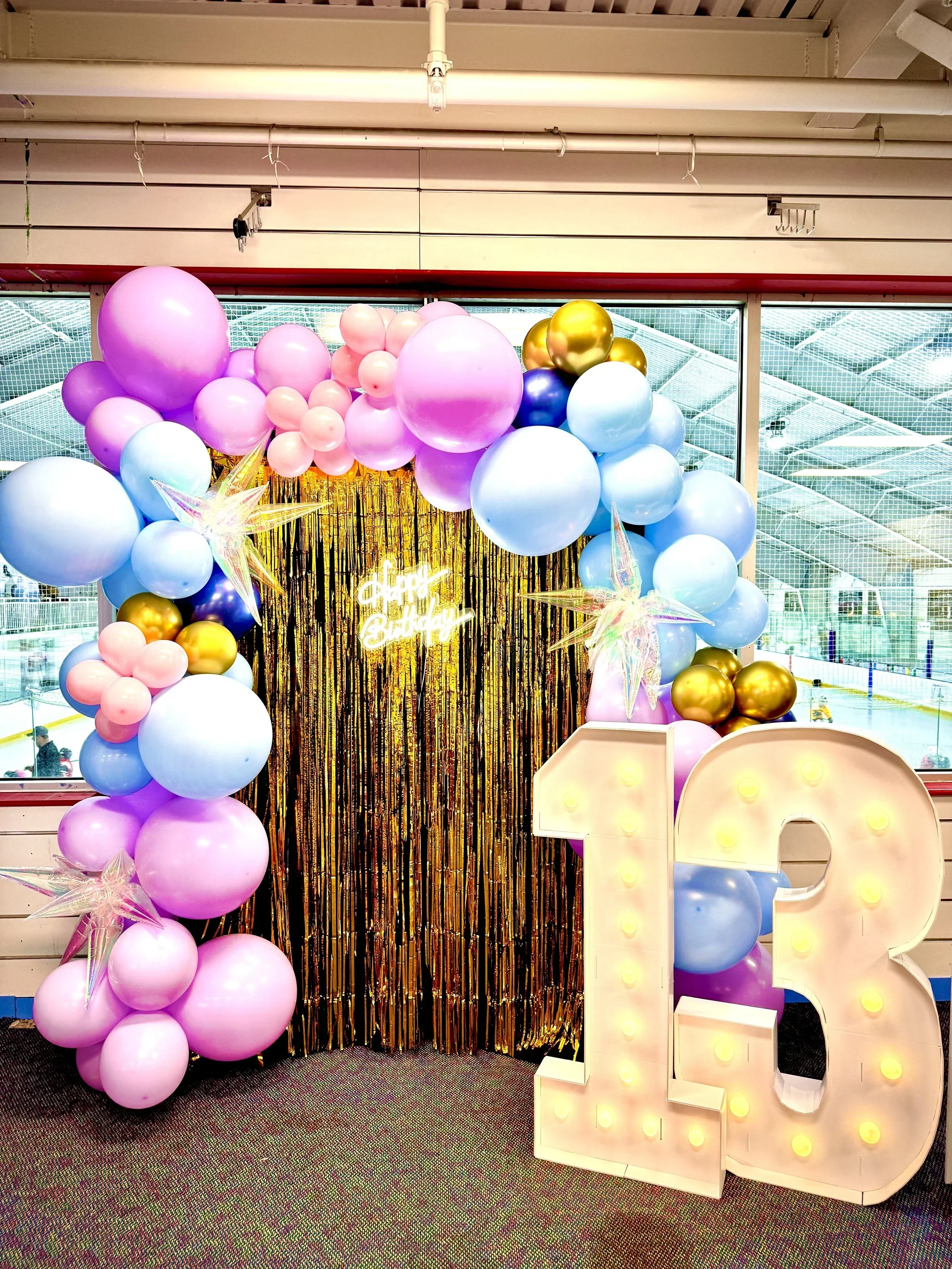 Decorative birthday celebration setup with pink, blue, gold, and purple balloons, a gold tinsel backdrop, illuminated marquee letter 'B', and a neon sign that says 'happy birthday' inside an ice rink area.